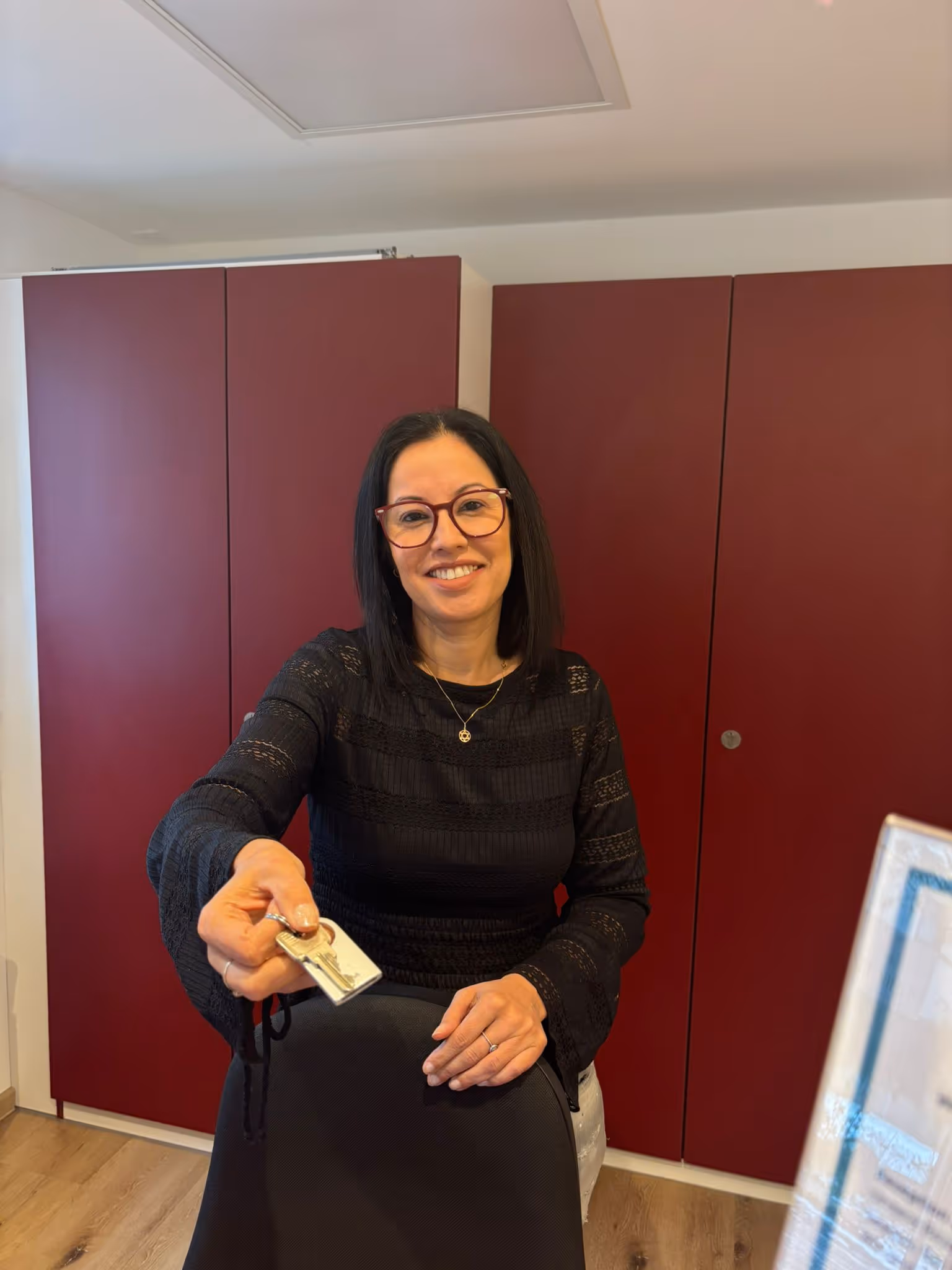 Smiling woman with glasses handing over a set of keys while sitting behind a black chair in a room with red cabinets.
