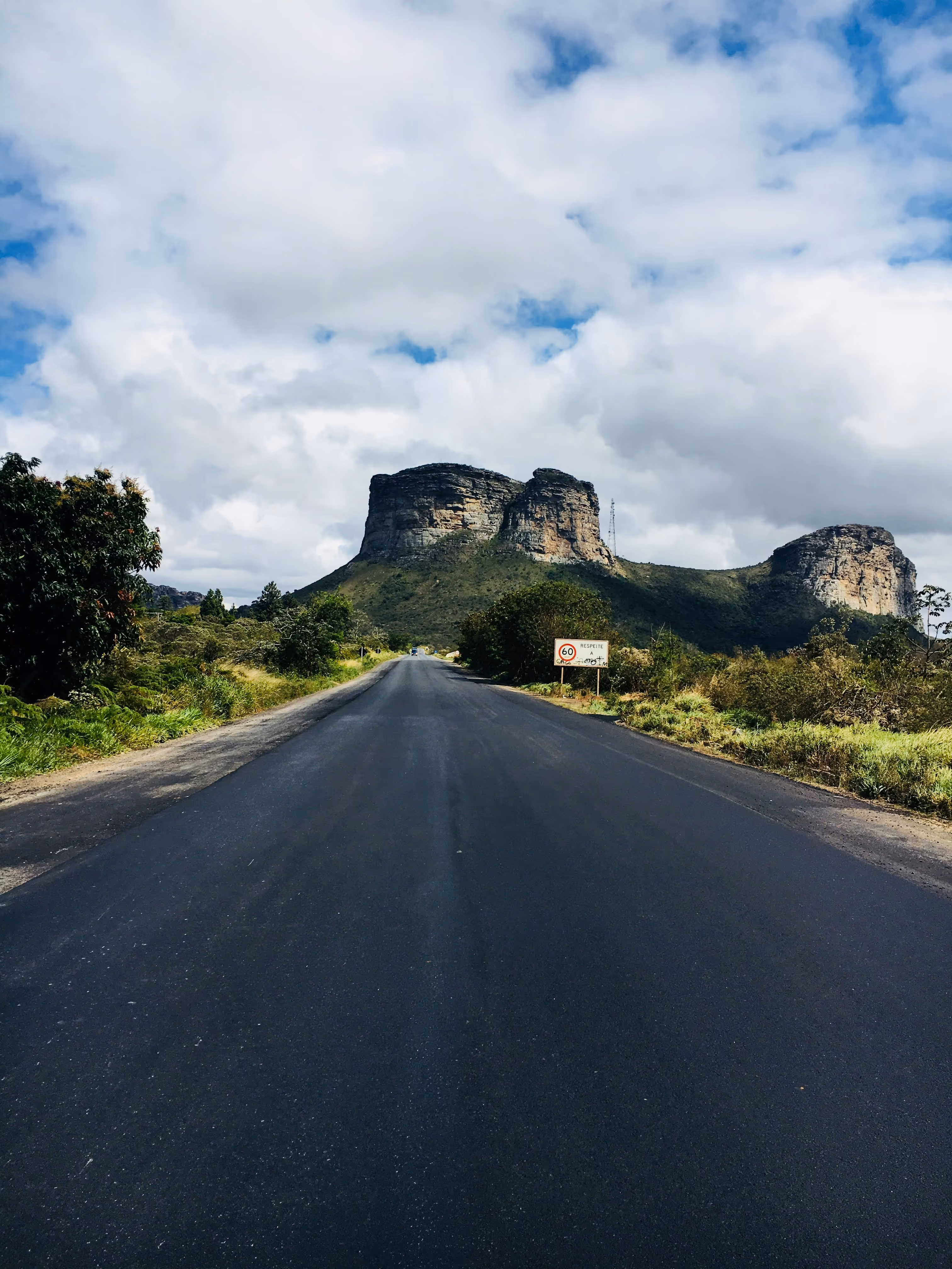 Straight asphalt road leading to a rocky hill with a partly cloudy sky overhead.