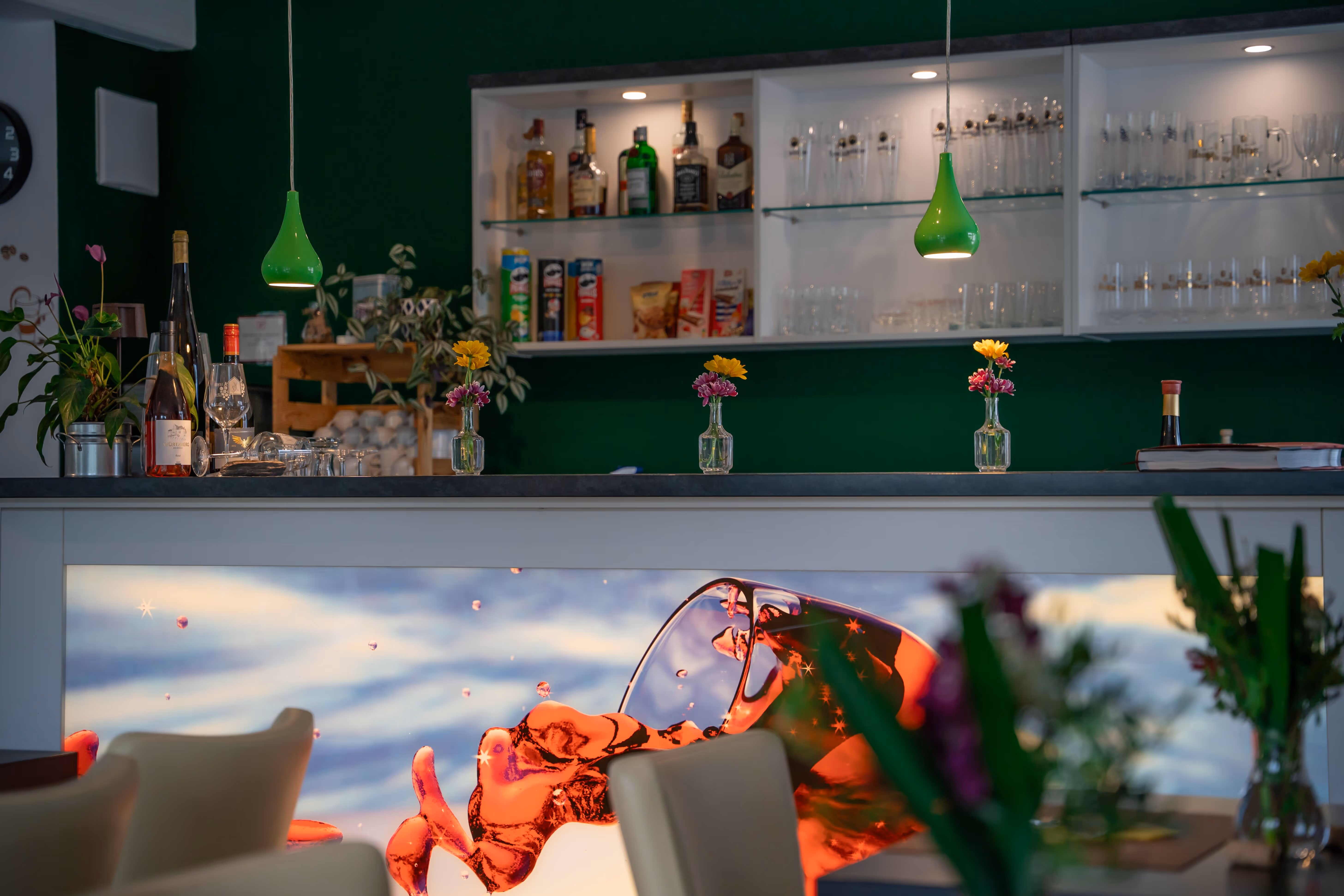 Modern bar counter with bottles, glassware, small flower vases, and green pendant lights against a dark green wall.