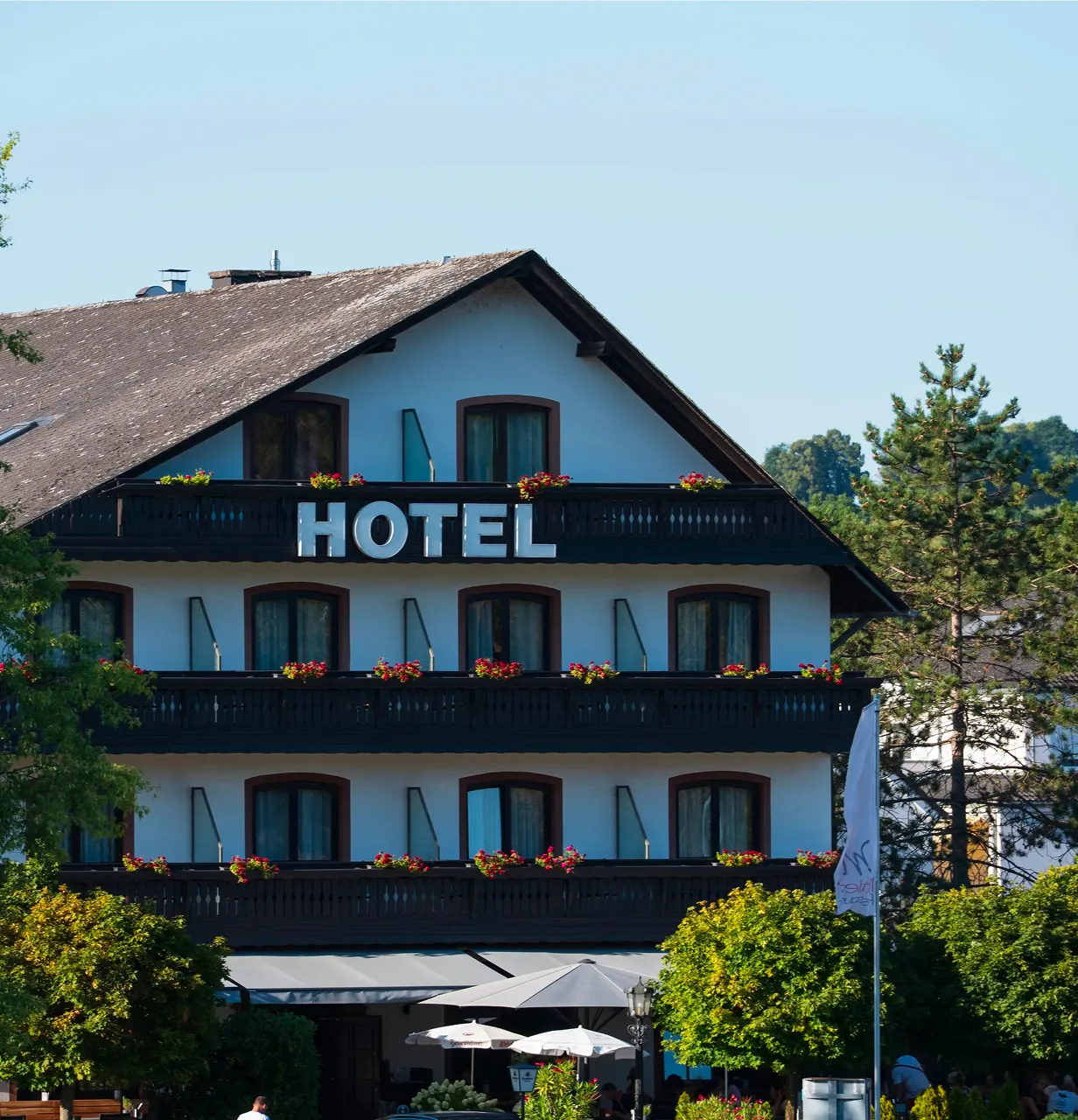 White multi-story hotel with balconies adorned with red flowers, surrounded by trees and greenery under a blue sky with scattered clouds.