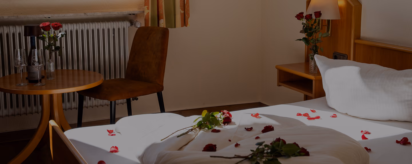 Hotel room bed with neatly folded towels, orange armchairs, a table with a wine bottle and flowers, and a bright wall lamp.