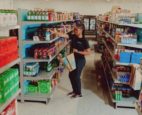 Woman with braided hair holding a shopping bag reaching for a drink on a grocery store shelf filled with beverages.