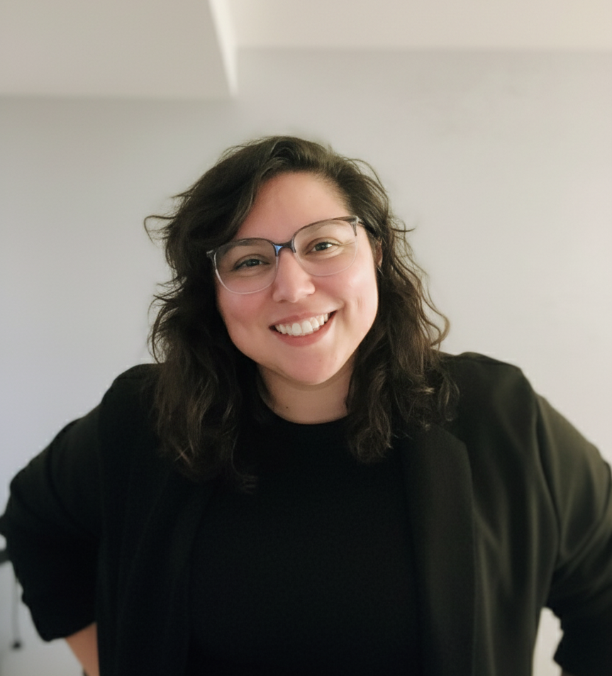 Smiling woman with wavy dark hair and glasses wearing a black top and jacket against a light background.