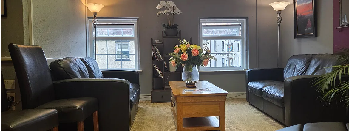 Cozy living room with black leather sofas, two chairs, wooden coffee table with flower vase, floor lamps, and two windows.