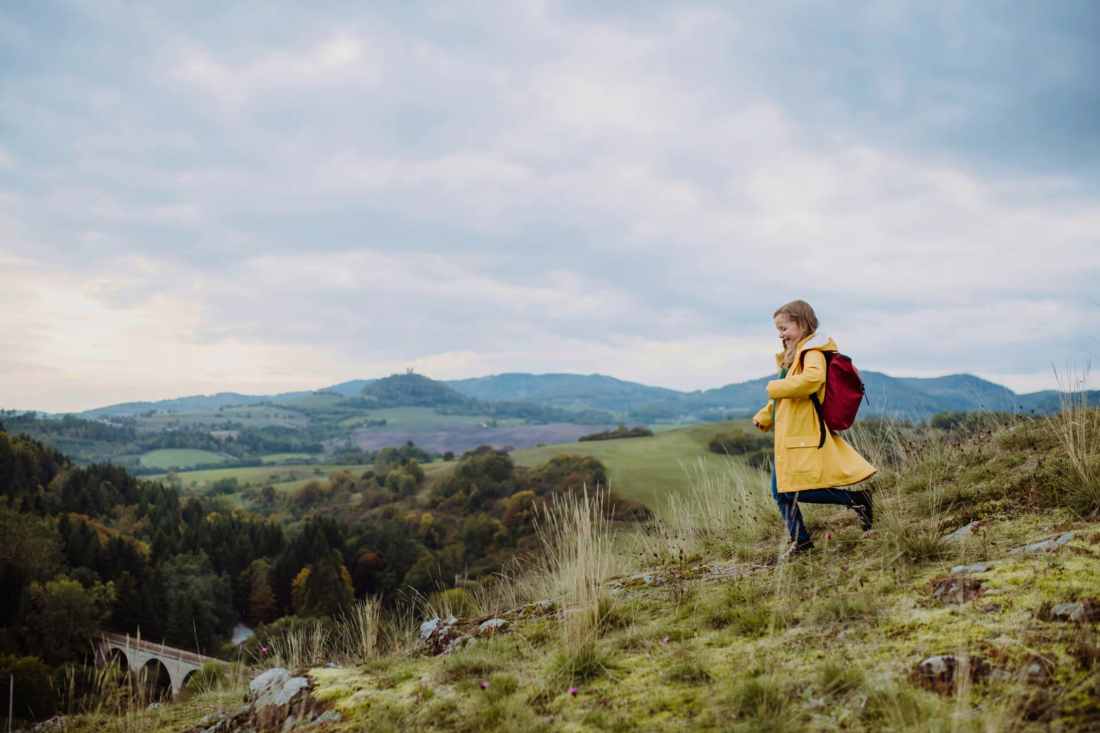 A young girl in a raincoat and backpack hikes through a grassy hillside with rolling valleys and distant mountains behind her.