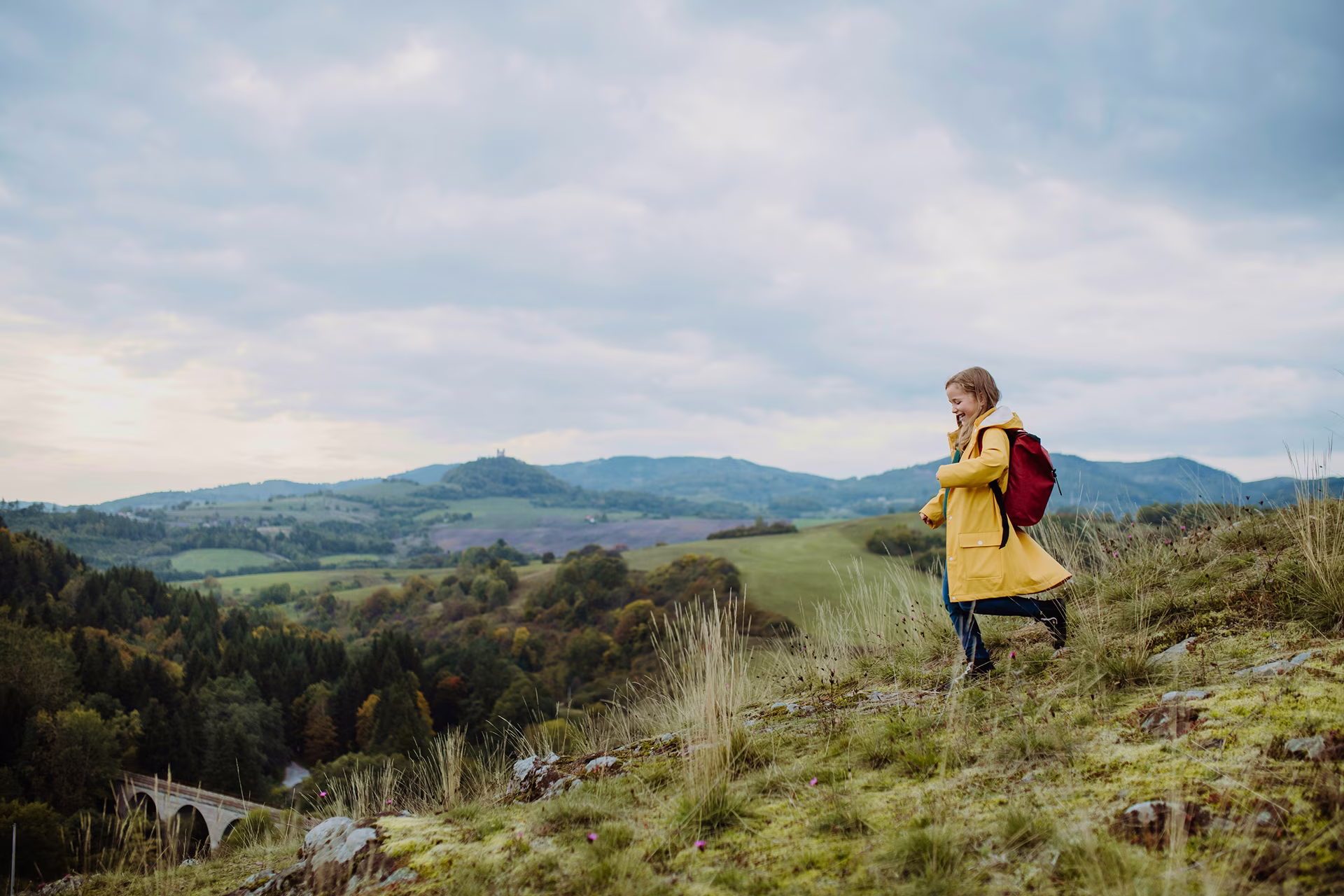 A young girl in a raincoat and backpack hikes through a grassy hillside with rolling valleys and distant mountains behind her.