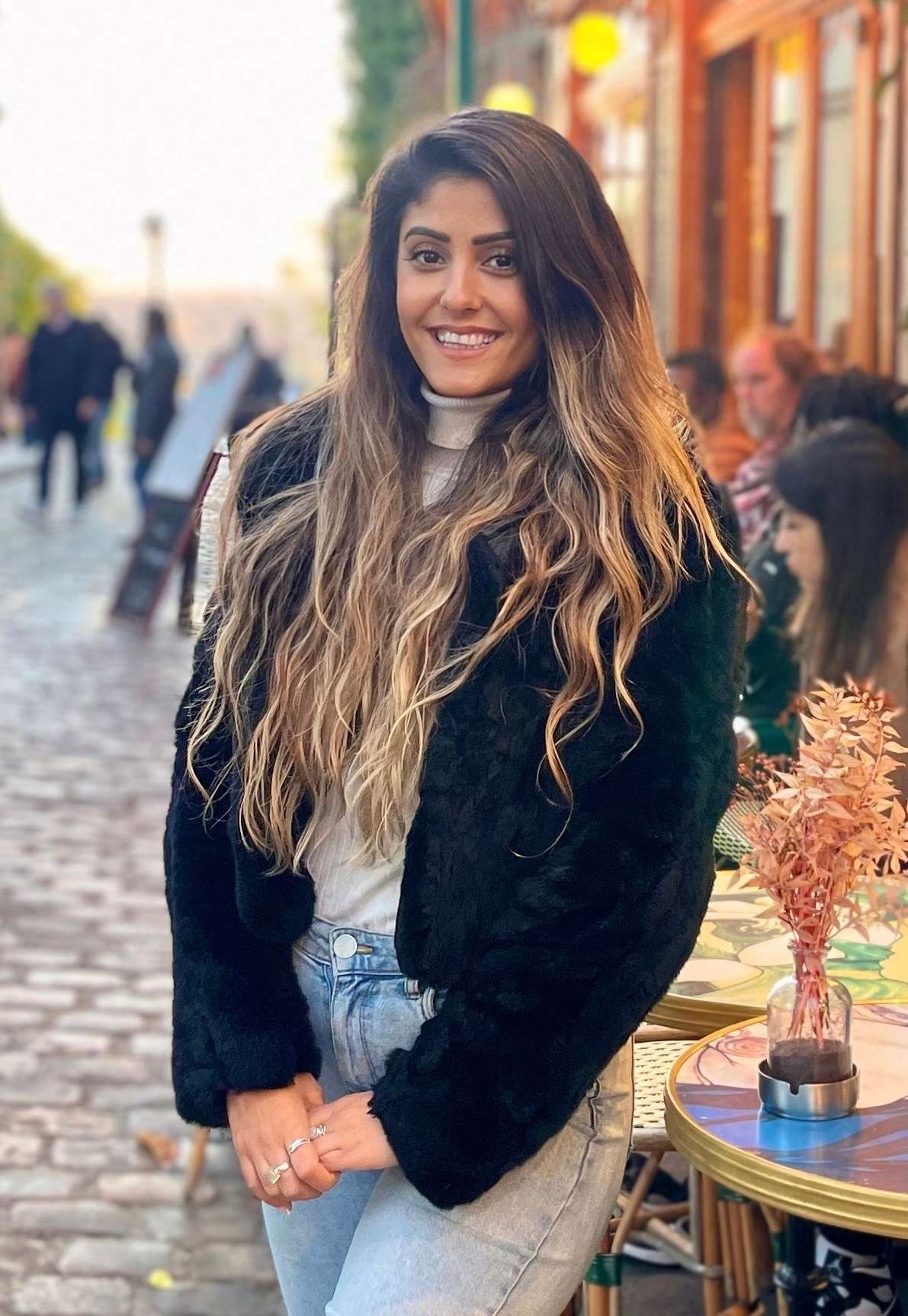 Young woman with long wavy hair wearing a black jacket and light blue jeans standing on a cobblestone street with outdoor cafe tables beside her.