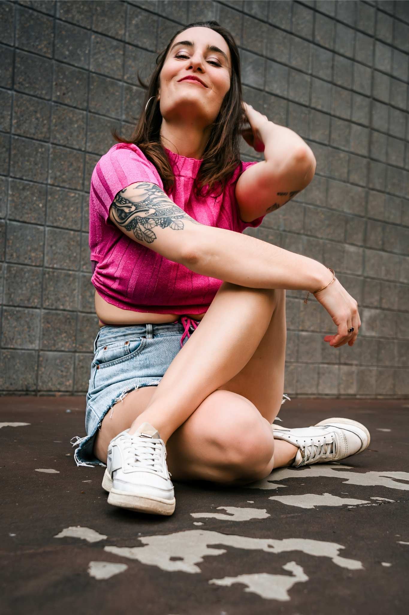 Woman with a tattoo on her arm wearing a pink top and denim shorts sitting cross-legged on a concrete surface against a tiled wall.