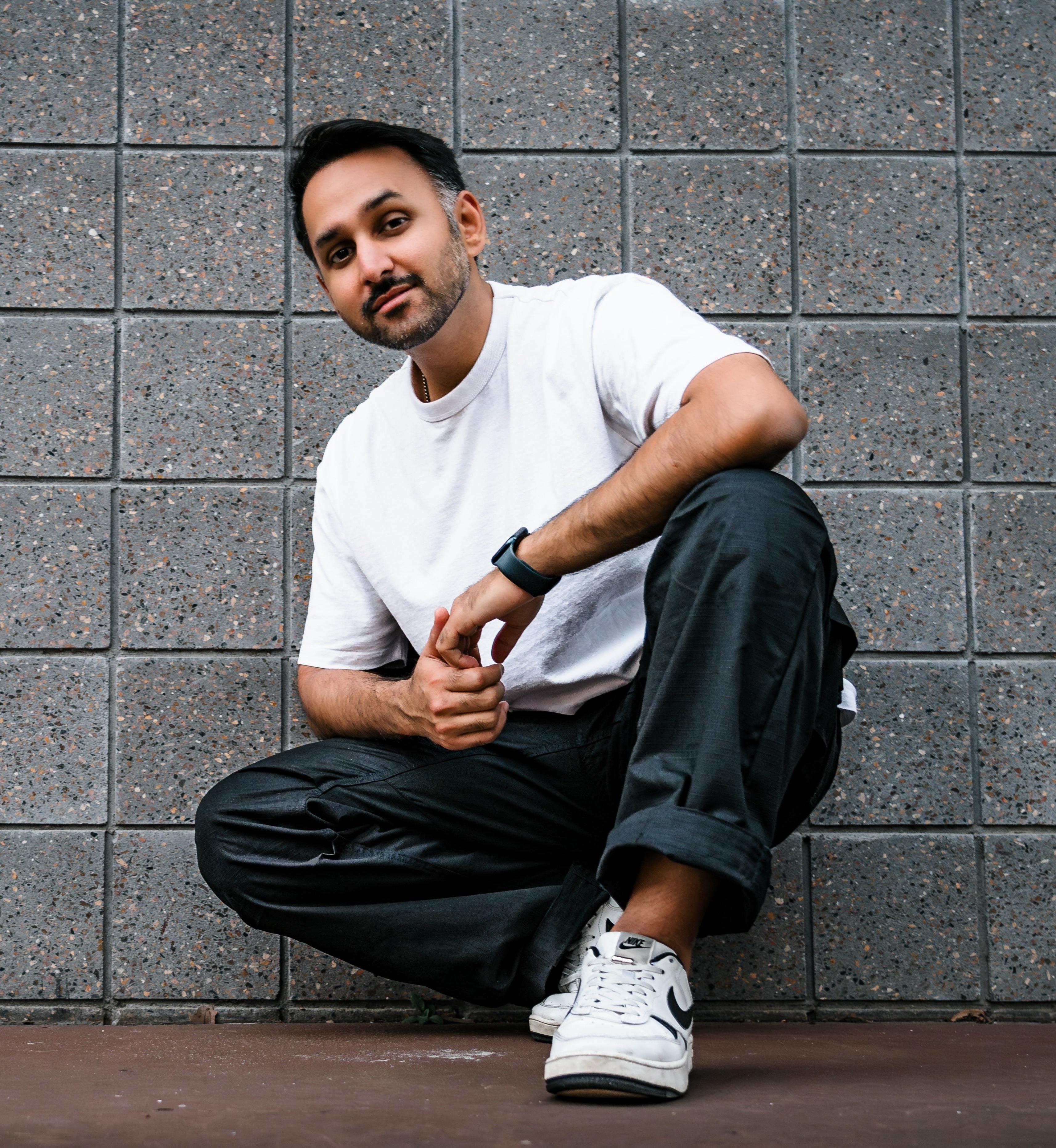 Man with beard wearing a white t-shirt and black pants crouching against a tiled gray wall.