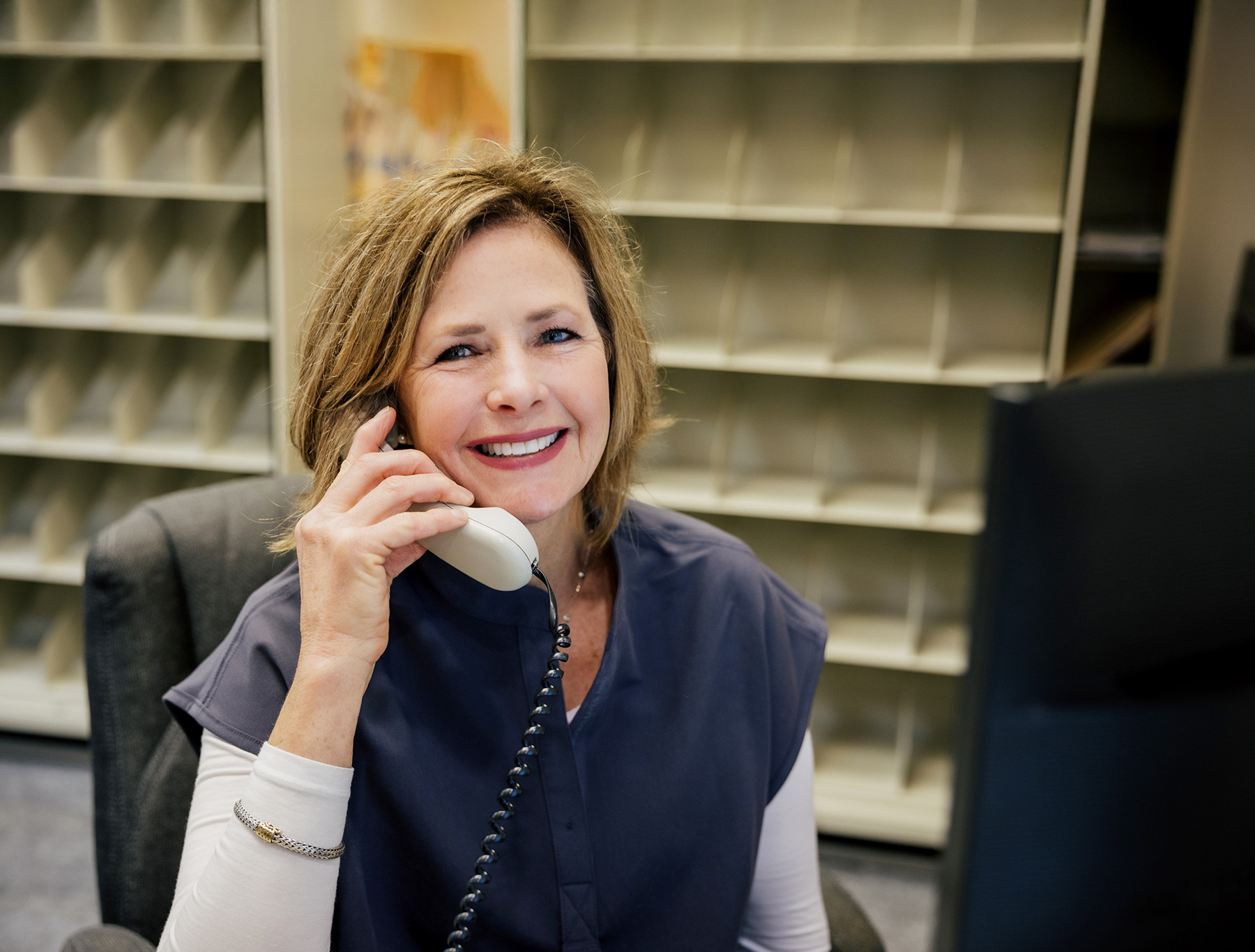 Smiling woman in a navy uniform talking on a corded phone at a desk with mail slots behind her.