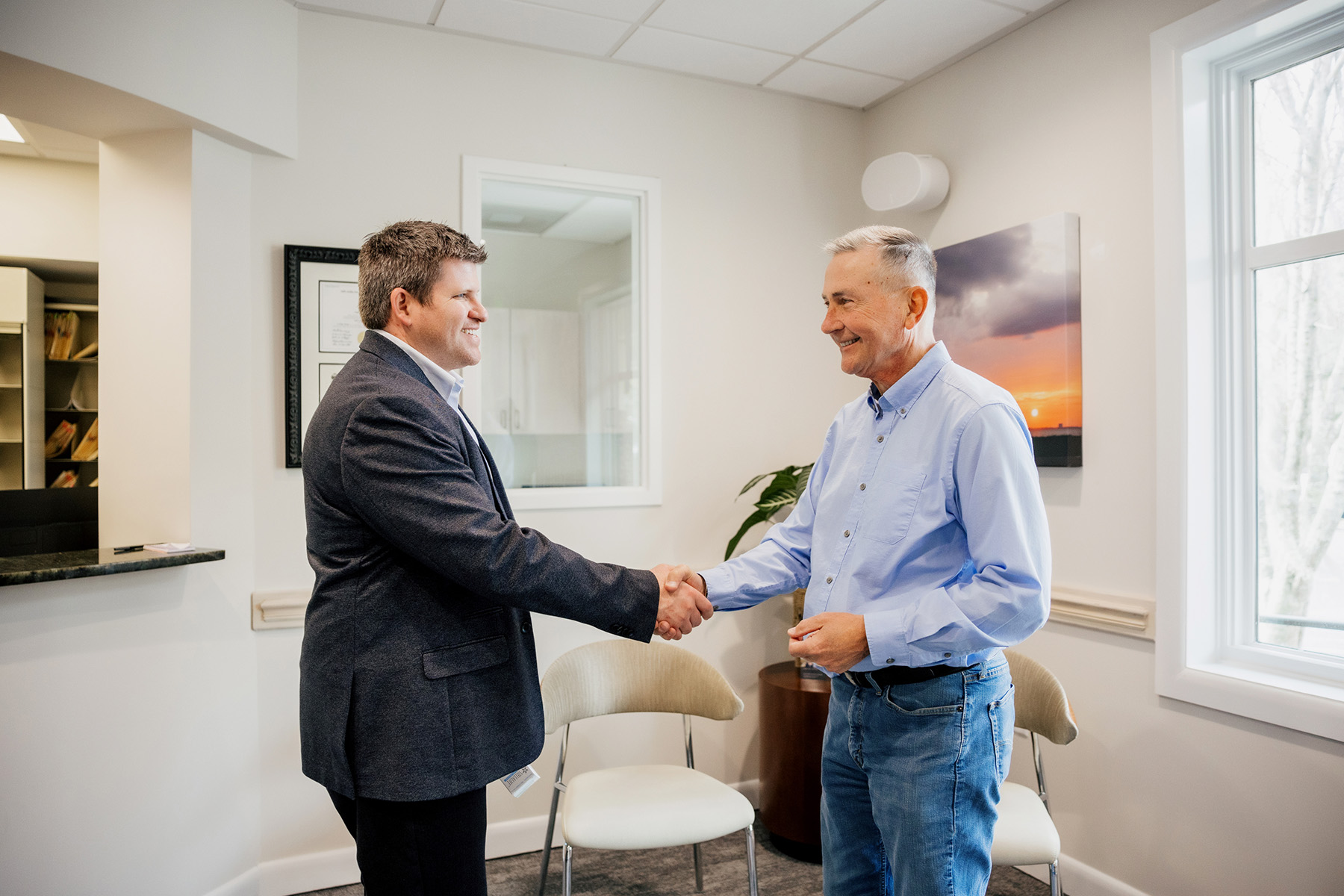 Two men smiling and shaking hands in a bright office with chairs, a plant, and a window showing a tree outside.