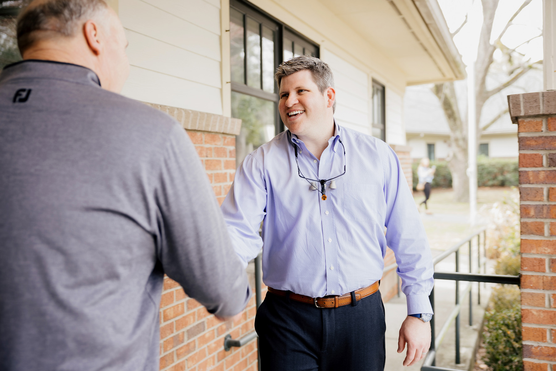 A man in a light blue shirt smiling as he shakes hands with another man wearing a gray shirt outside a brick building.
