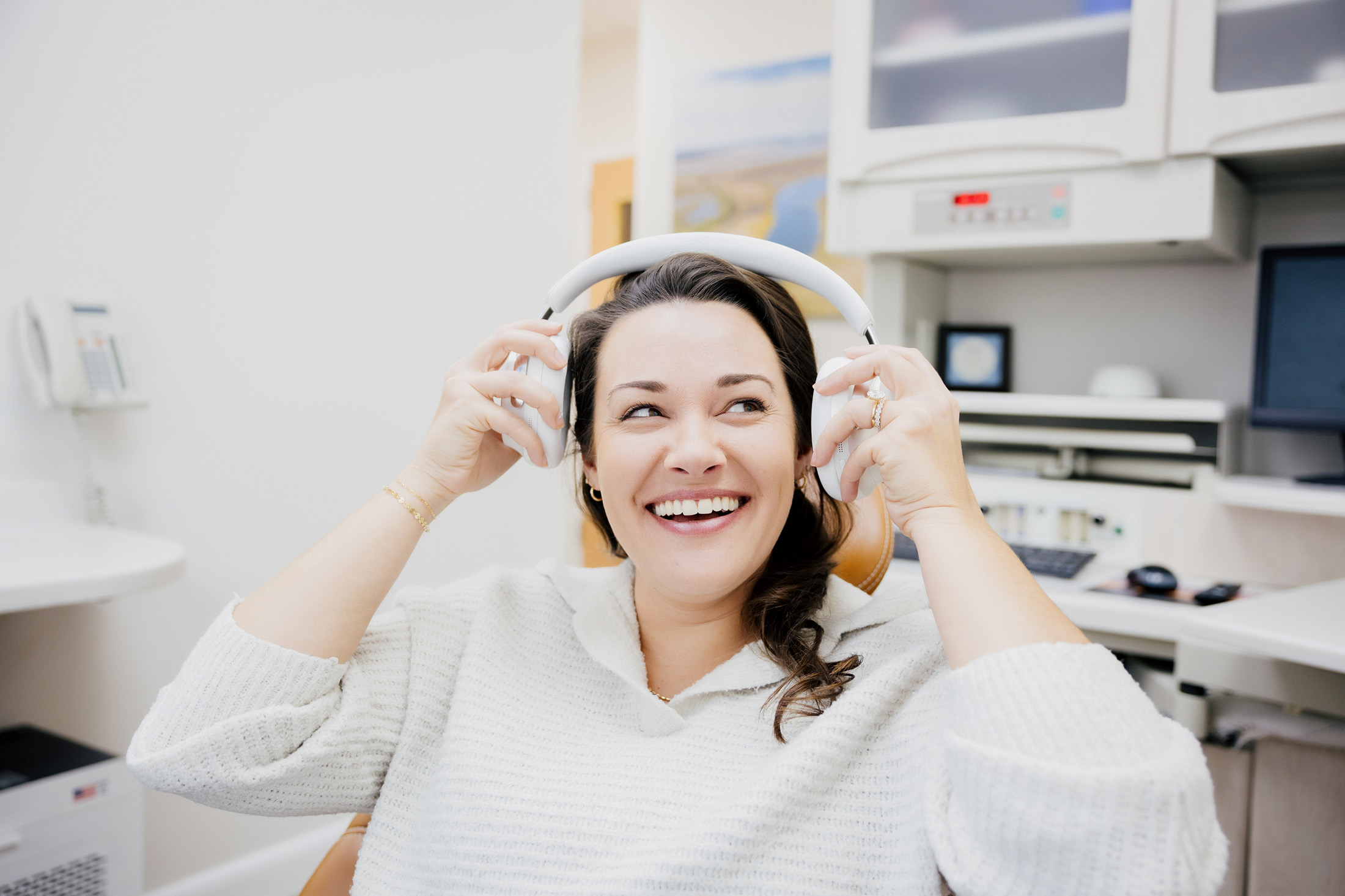 Smiling woman wearing a white sweater putting on white over-ear headphones in a modern office setting.