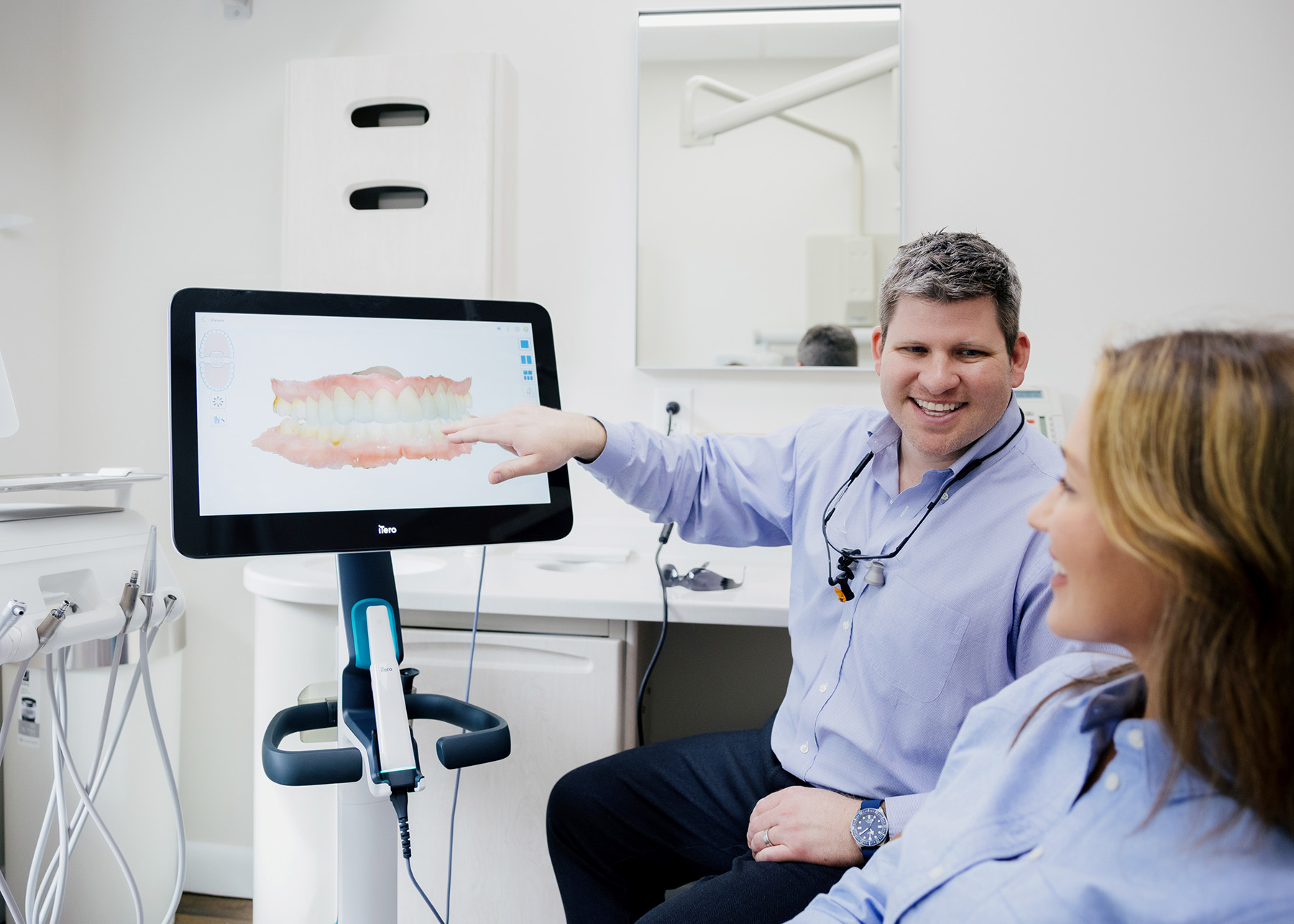 Dentist explaining a dental scan image on a digital screen to a smiling female patient in a dental office.