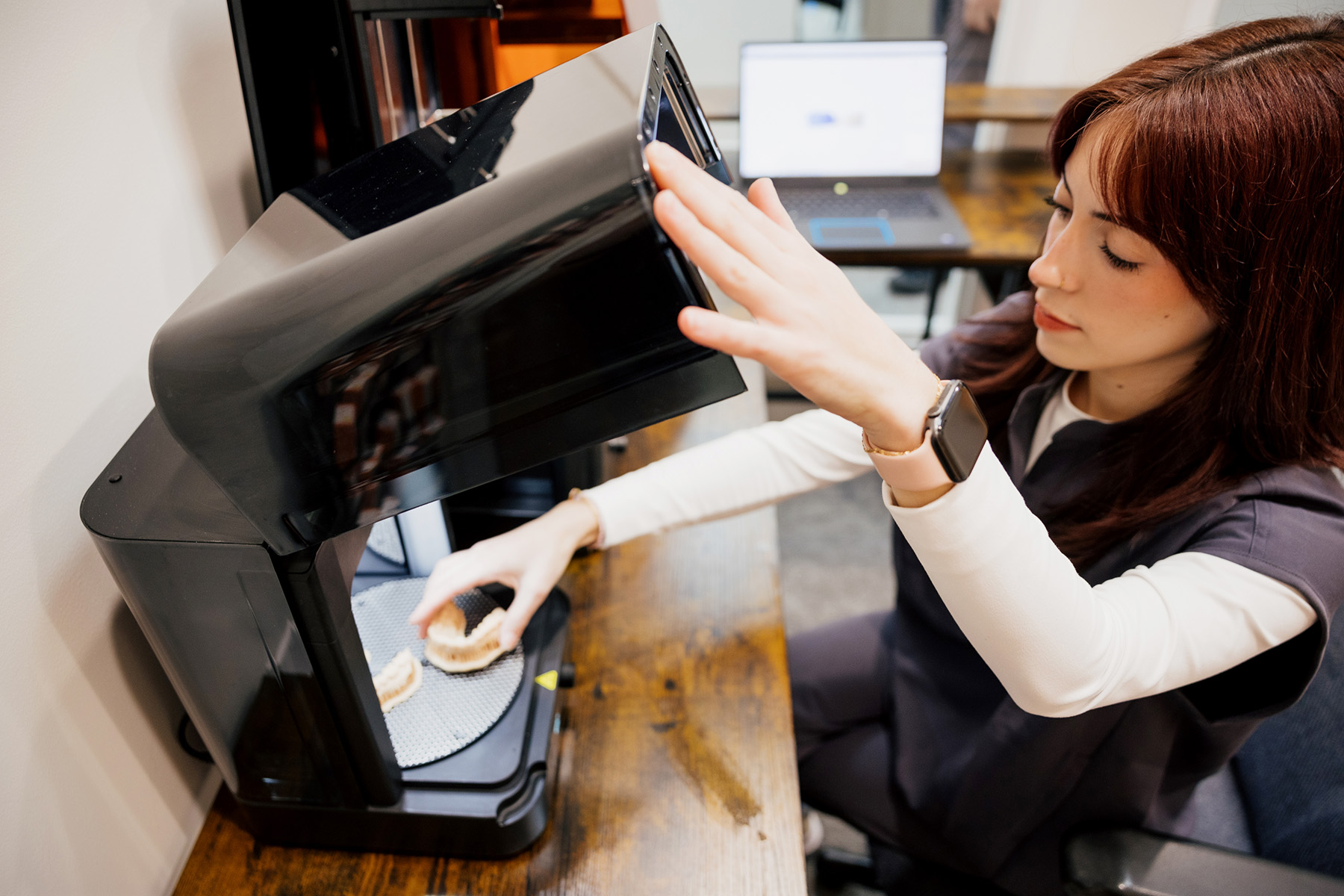 Woman placing dental molds inside a 3D scanner on a wooden desk with a laptop in the background.