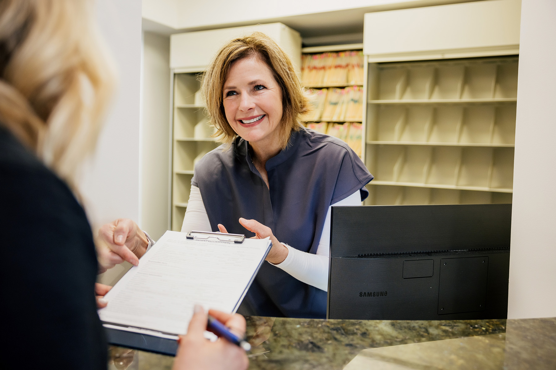 Smiling receptionist helping a patient with a clipboard at a medical office front desk.
