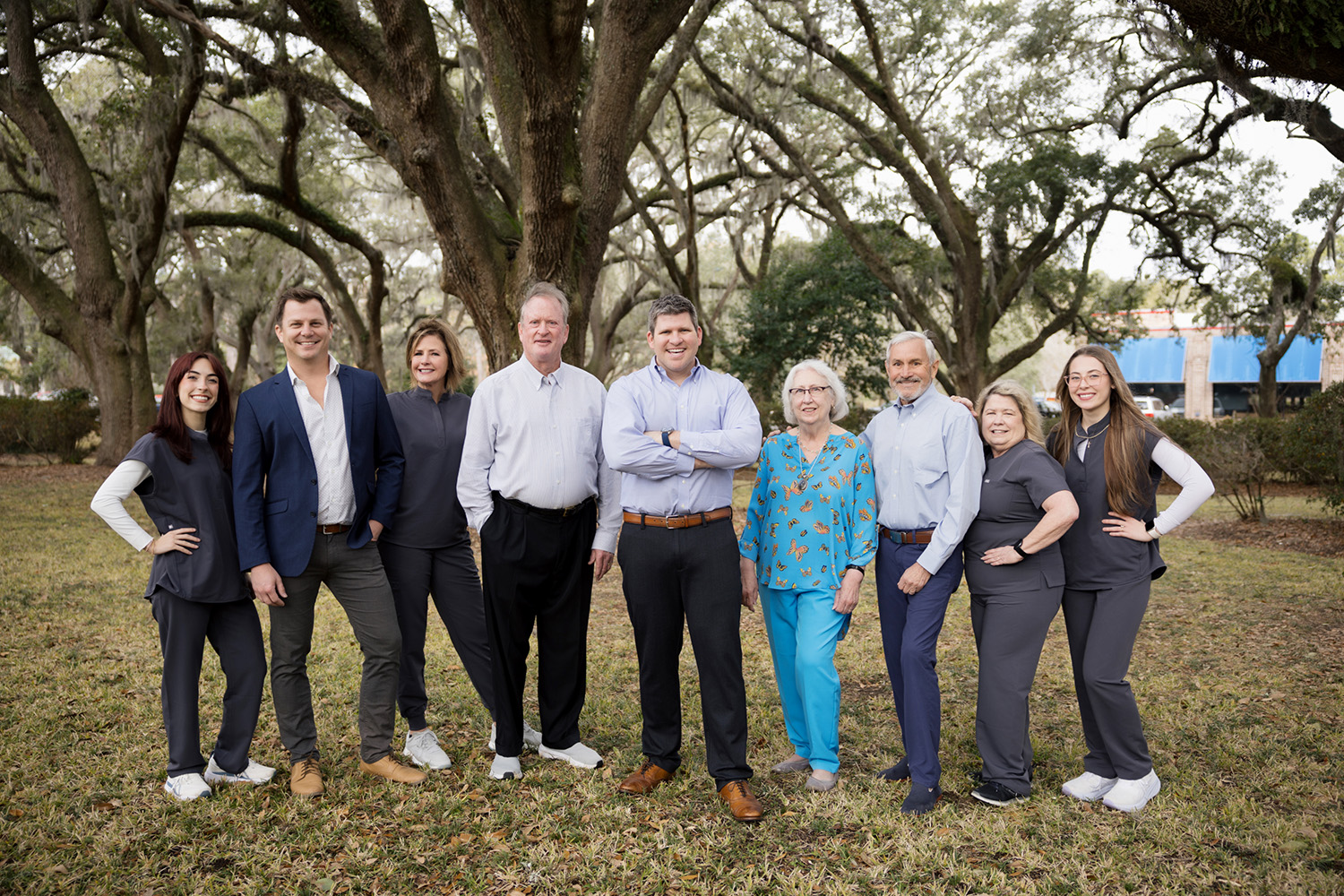 Group of nine adults standing outdoors on grass with large trees in the background, smiling at the camera.