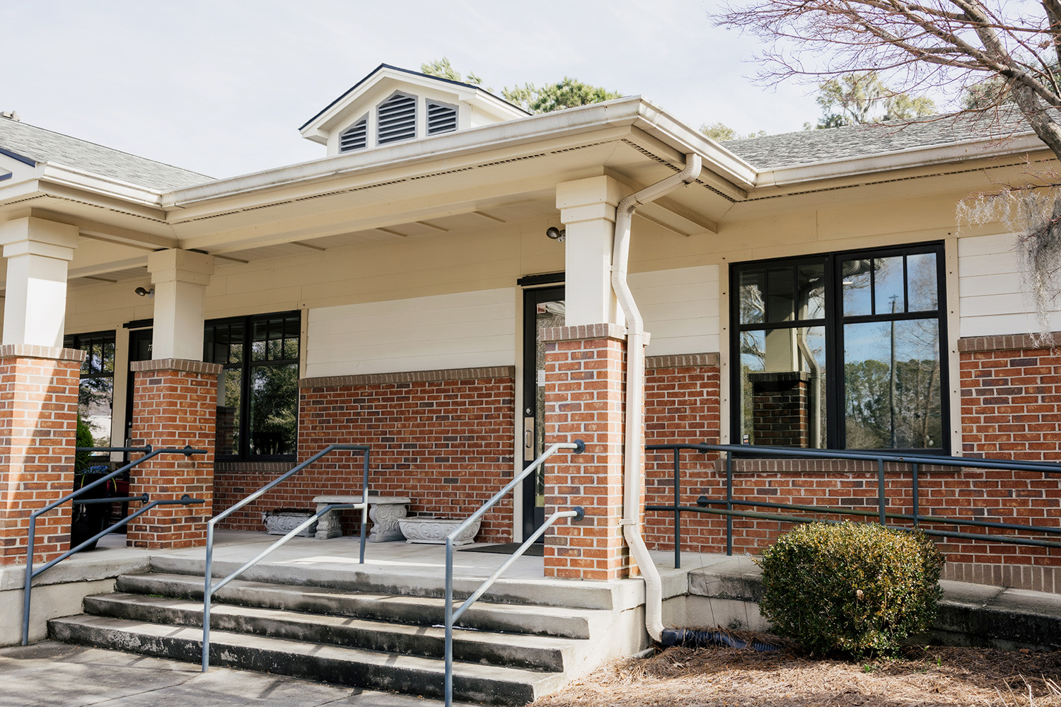 Entrance of a brick building with concrete steps, metal railings, and a ramp leading to a black door under a covered porch.