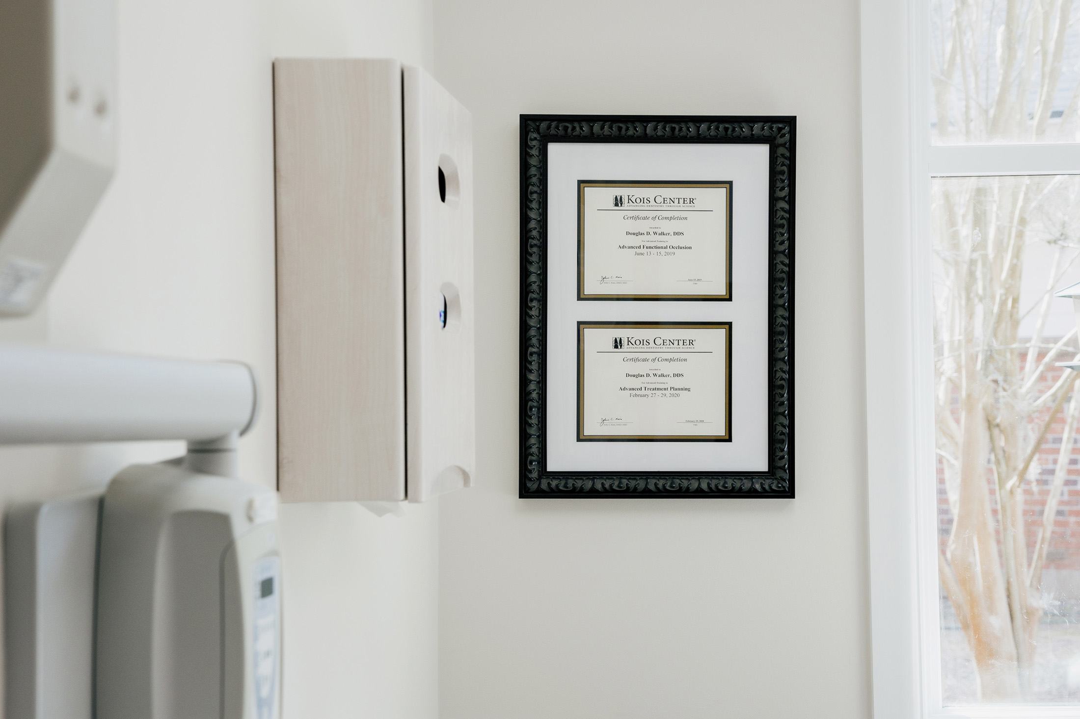 Two framed certificates from Kois Center for Douglas D. Walker, DDS, hanging on a white wall near a window.