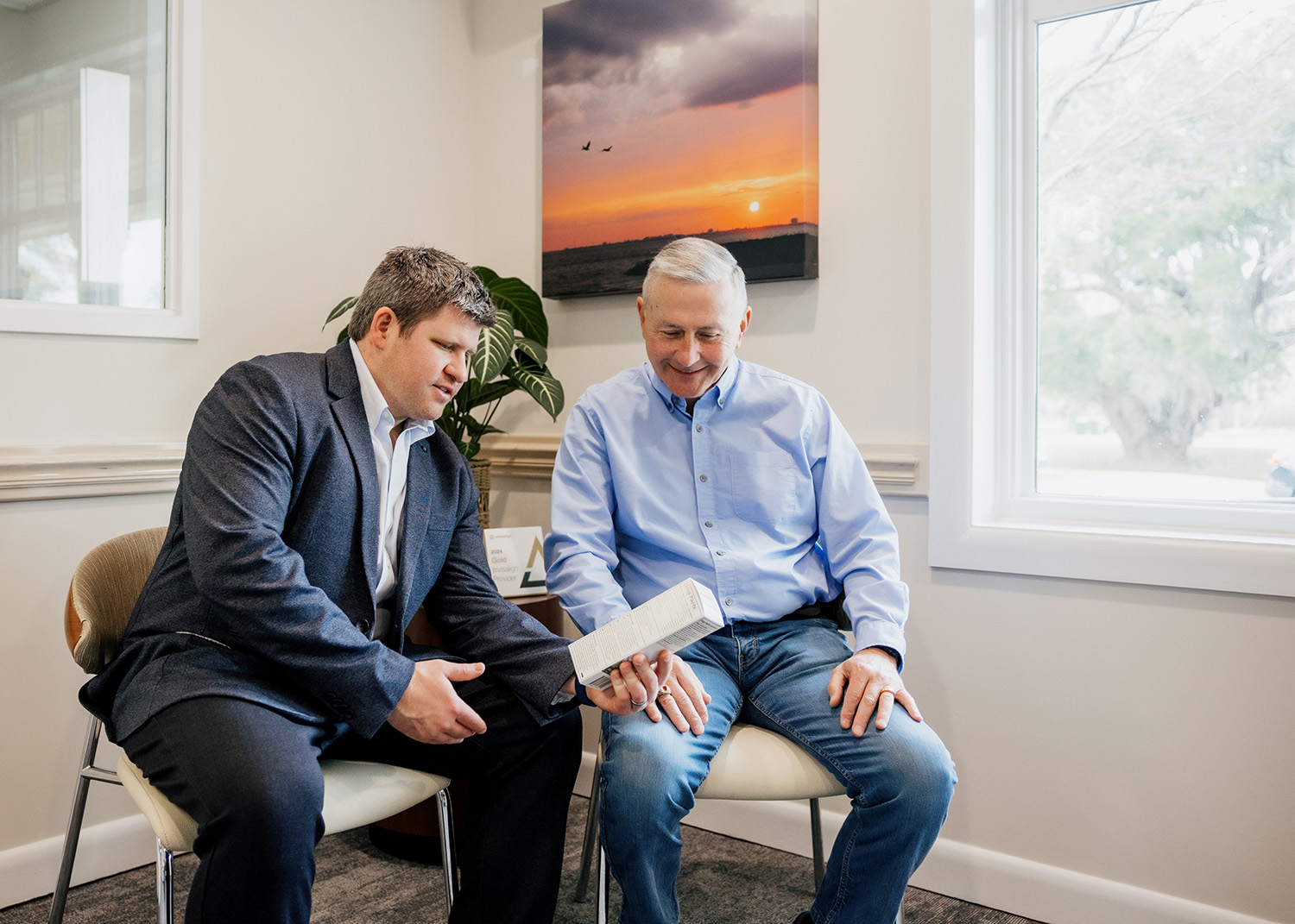 Two men seated indoors, one in a suit explaining a product on a box to an older man in a light blue shirt.