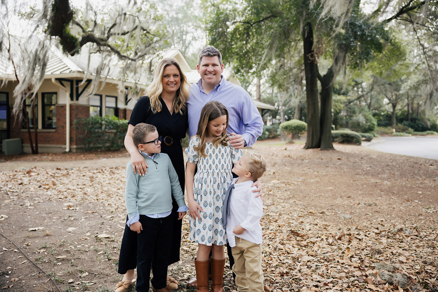 Smiling family of five standing outdoors on fallen leaves with trees and a brick building in the background.