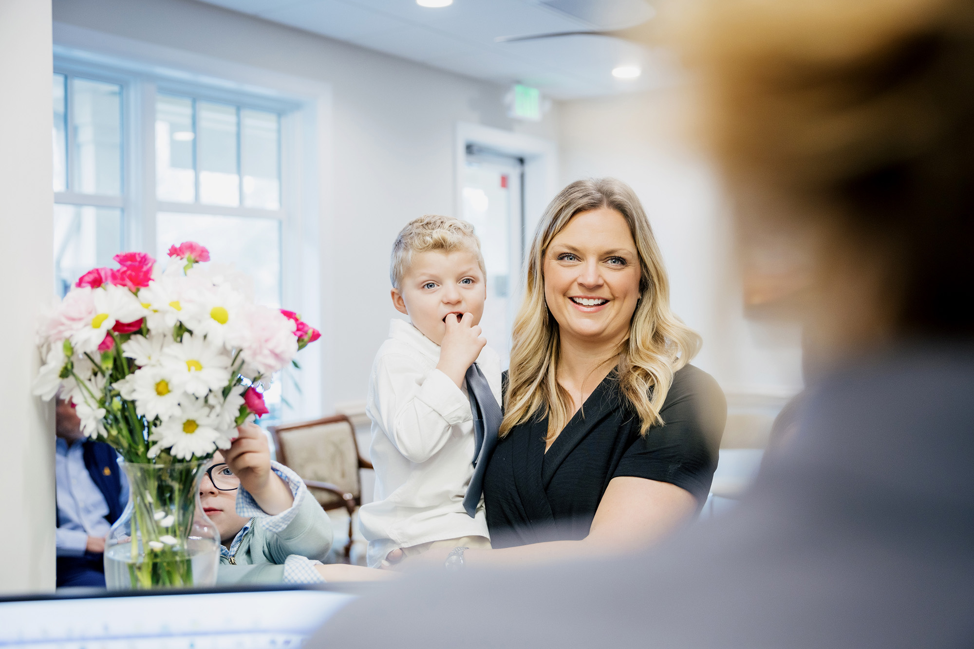 A smiling woman holding a young boy with a tie while another child reaches for flowers in a vase in a bright room.