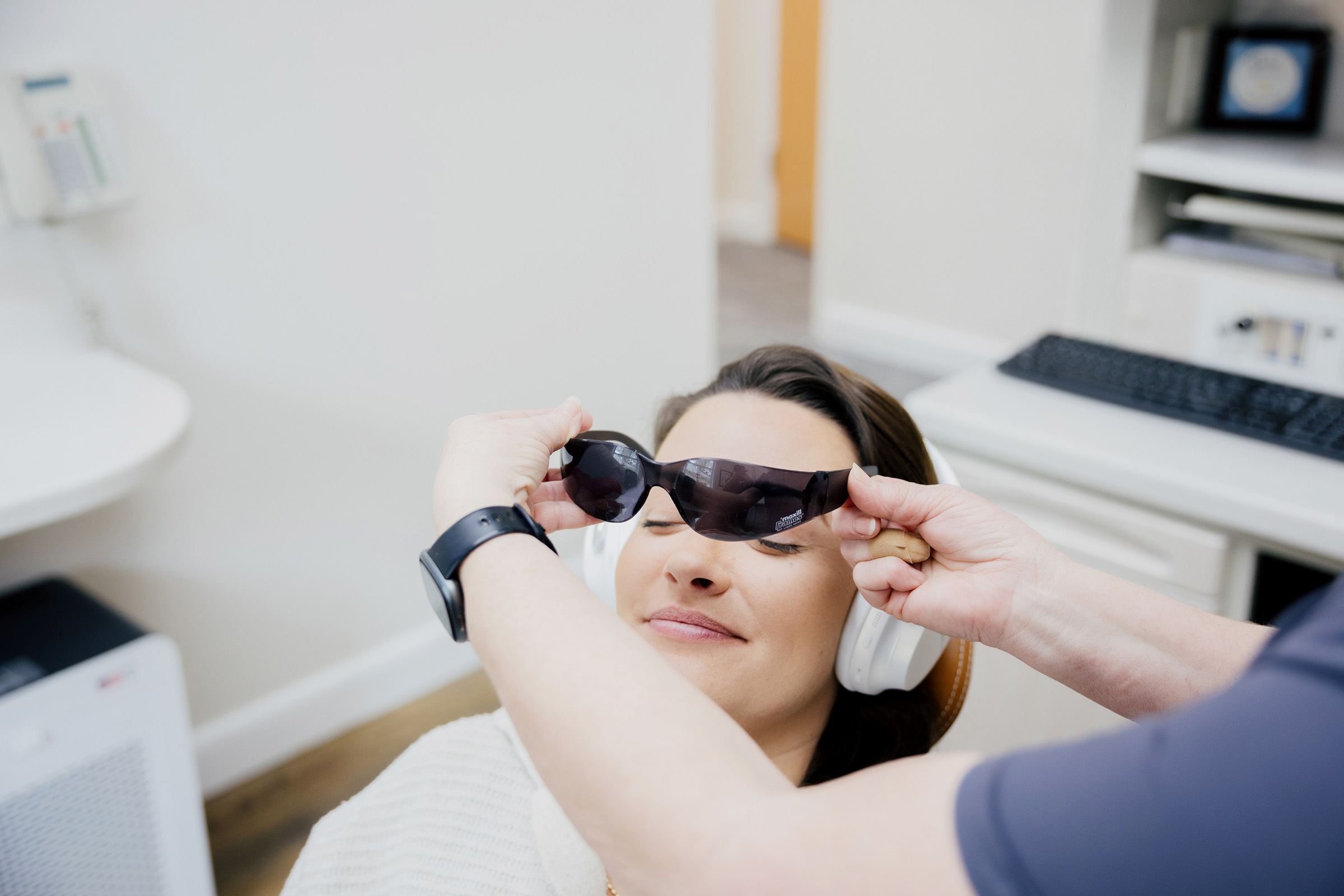 A woman with headphones sits in a clinic chair as a person holds a pair of dark protective glasses in front of her eyes.