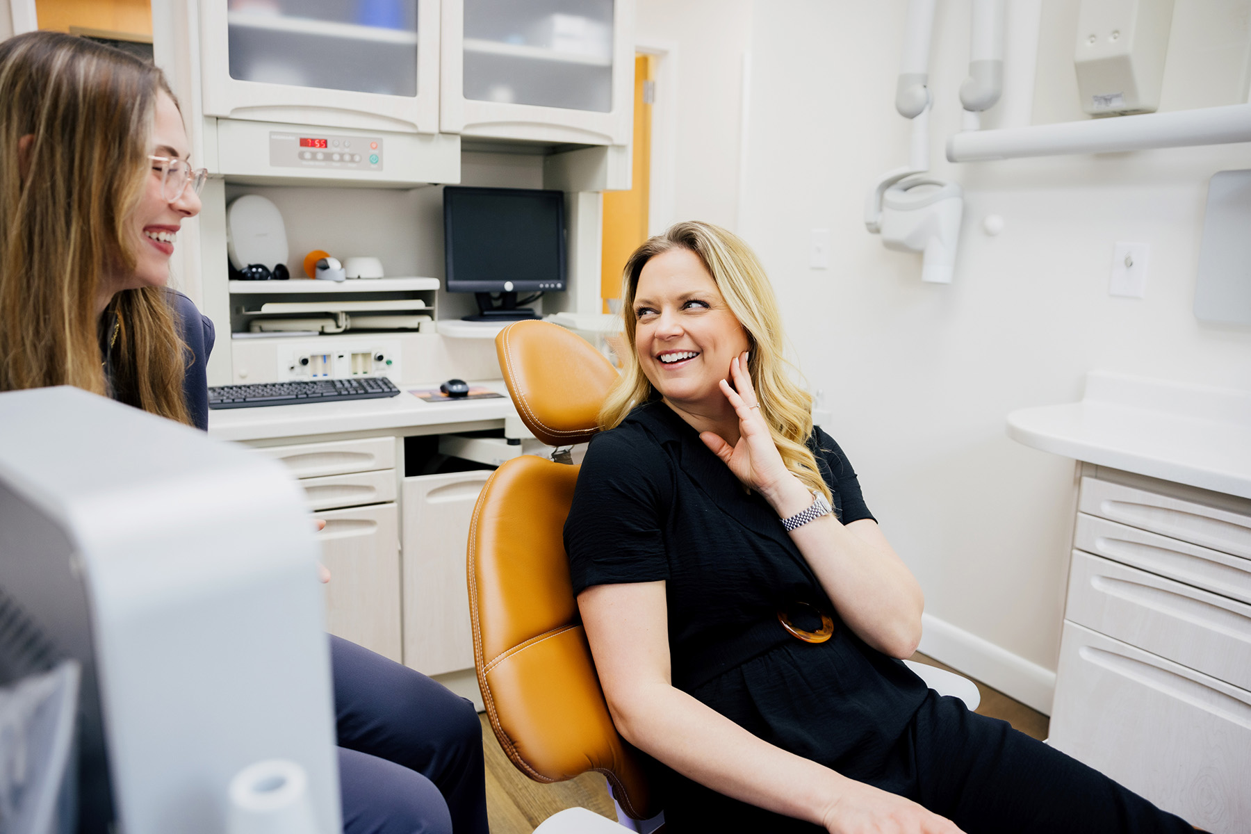 Smiling woman sitting in a dental chair talking to a female dentist in a bright dental office.