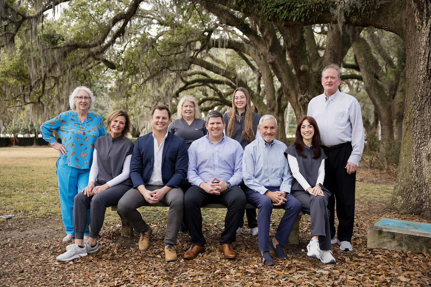 A group of ten adults posing outdoors on a wooden bench under large oak trees with Spanish moss.