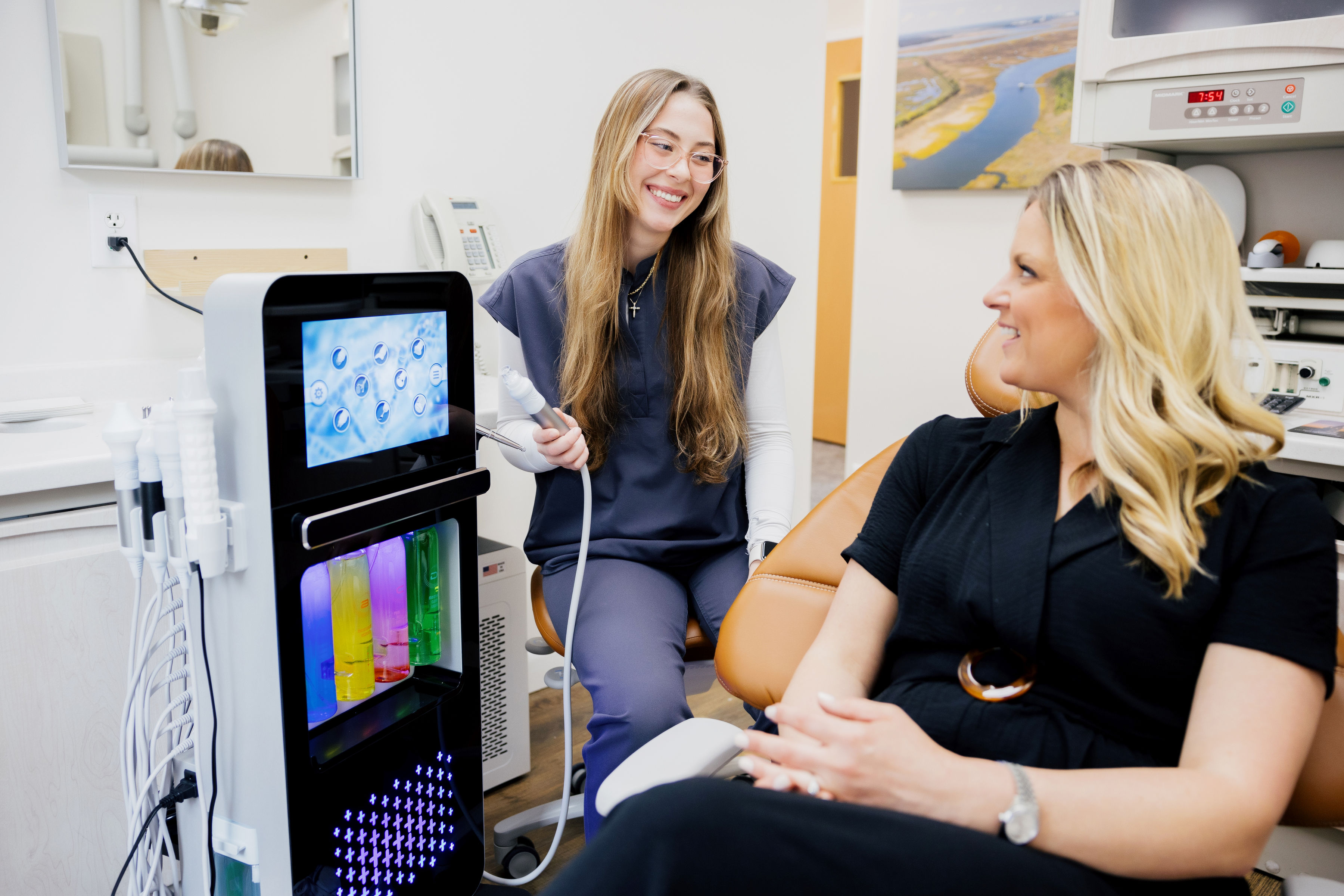 Smiling dental hygienist holding a dental tool while talking to a female patient sitting in a dental chair.