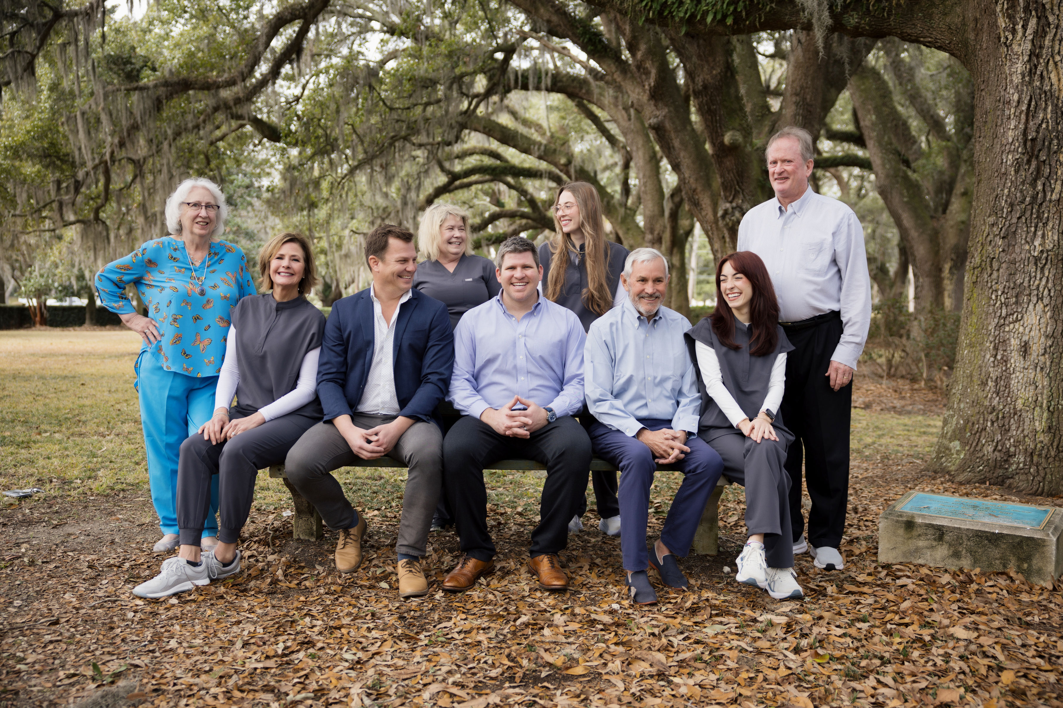 Group of nine adults smiling and sitting and standing outdoors on a bench under large trees with hanging moss.