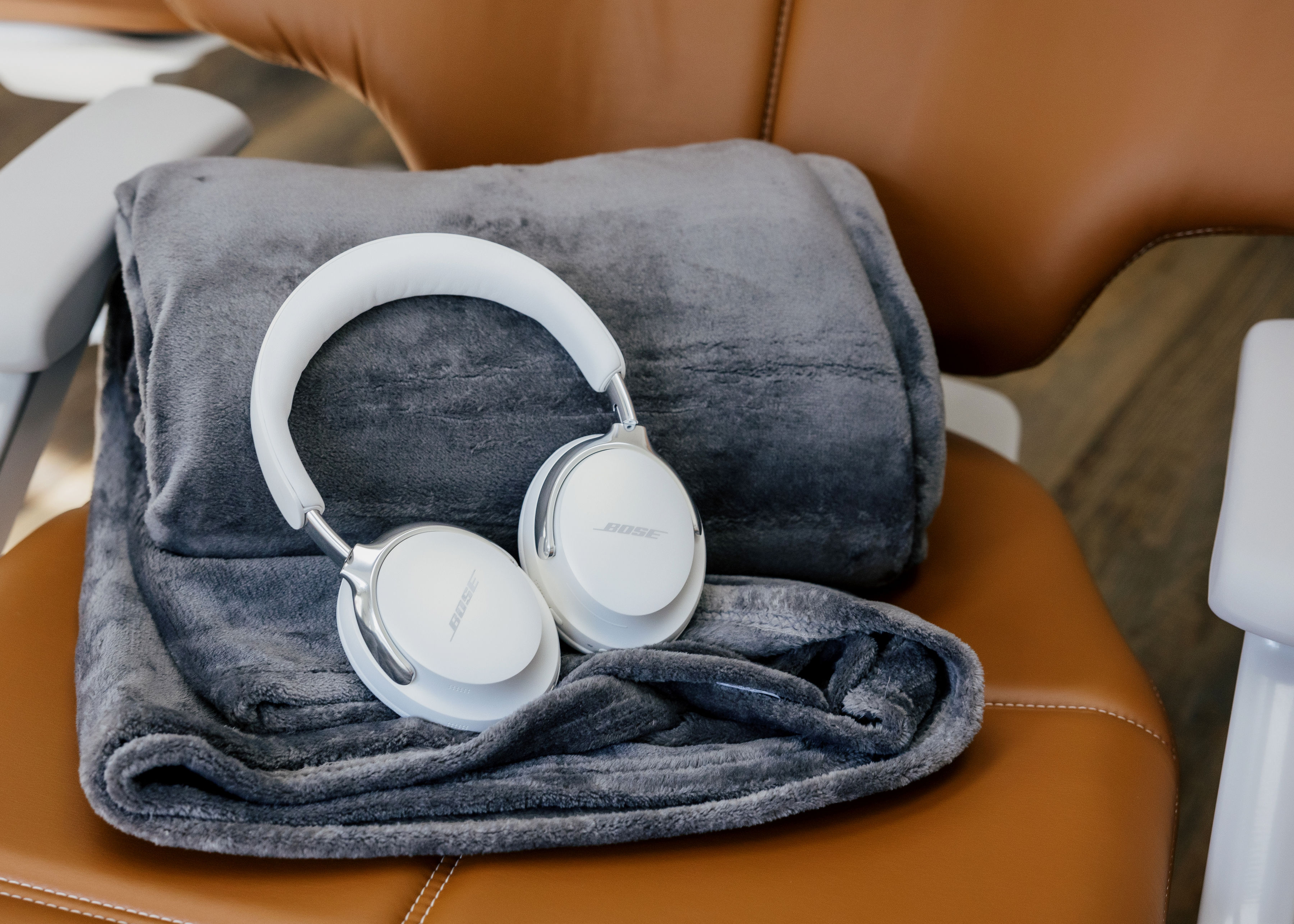 White Bose over-ear headphones resting on a folded gray blanket on a brown leather chair.