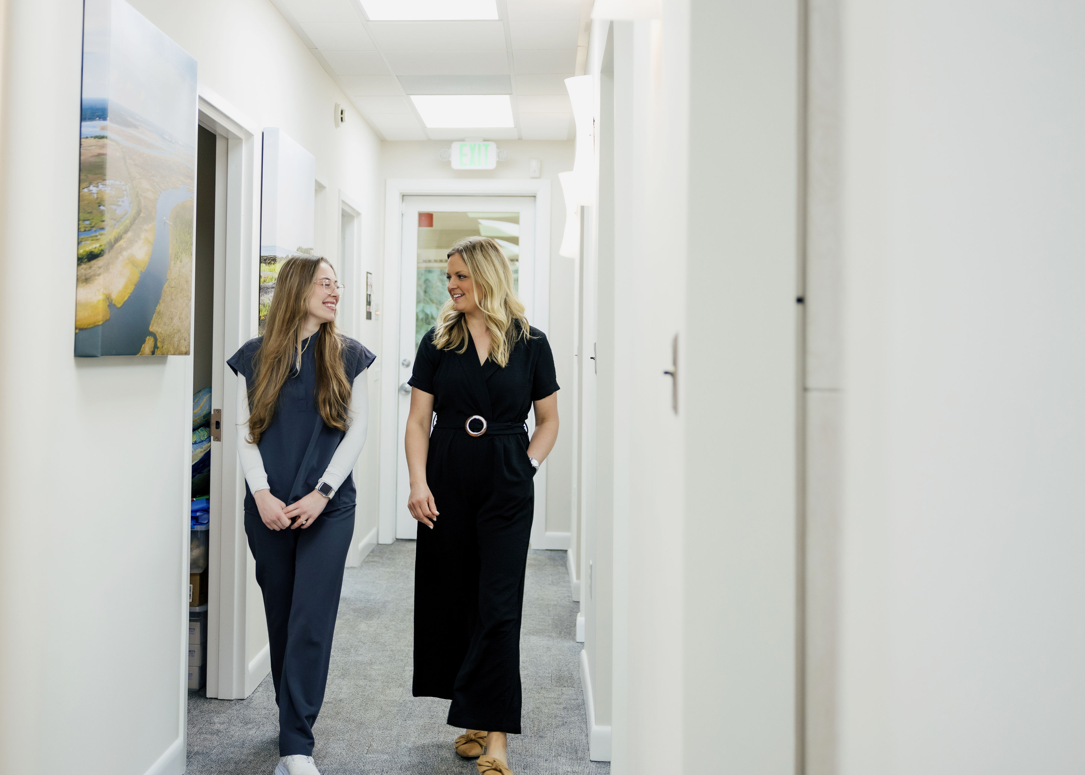 Two women walking and smiling in a bright hallway with white walls and gray carpet.