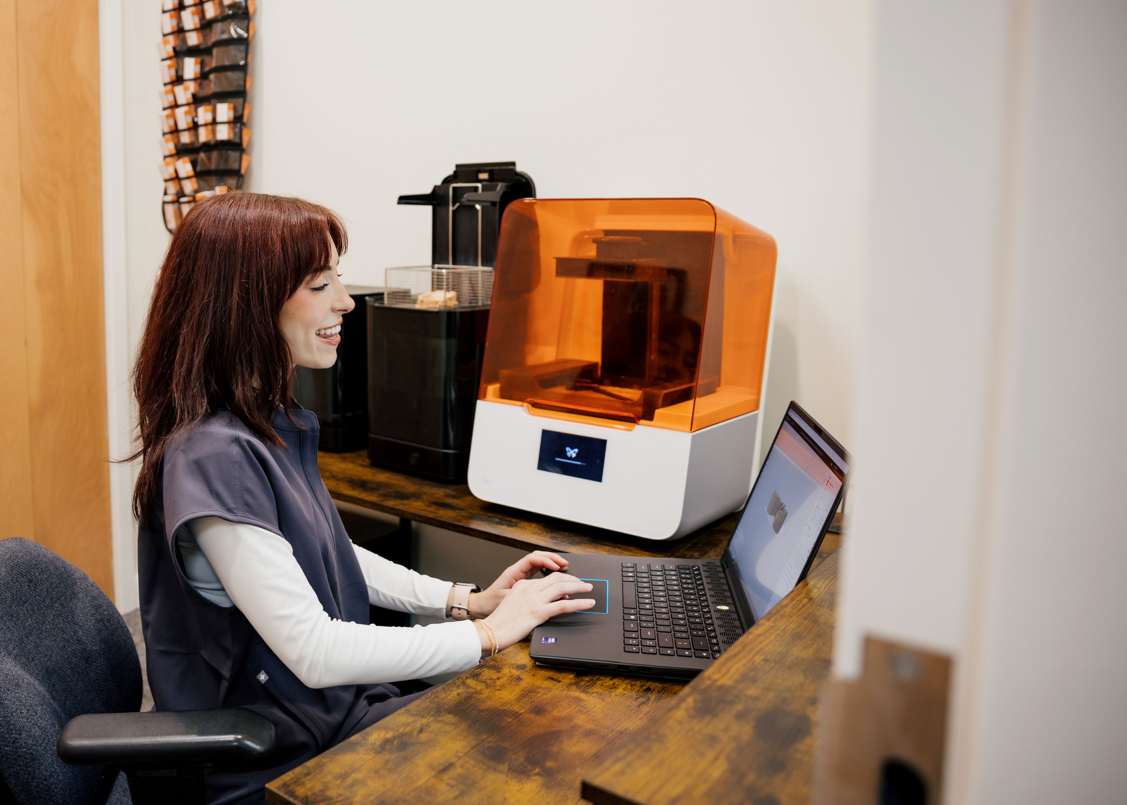 Smiling woman in work attire sitting at a wooden desk using a laptop beside an orange-covered 3D printer.