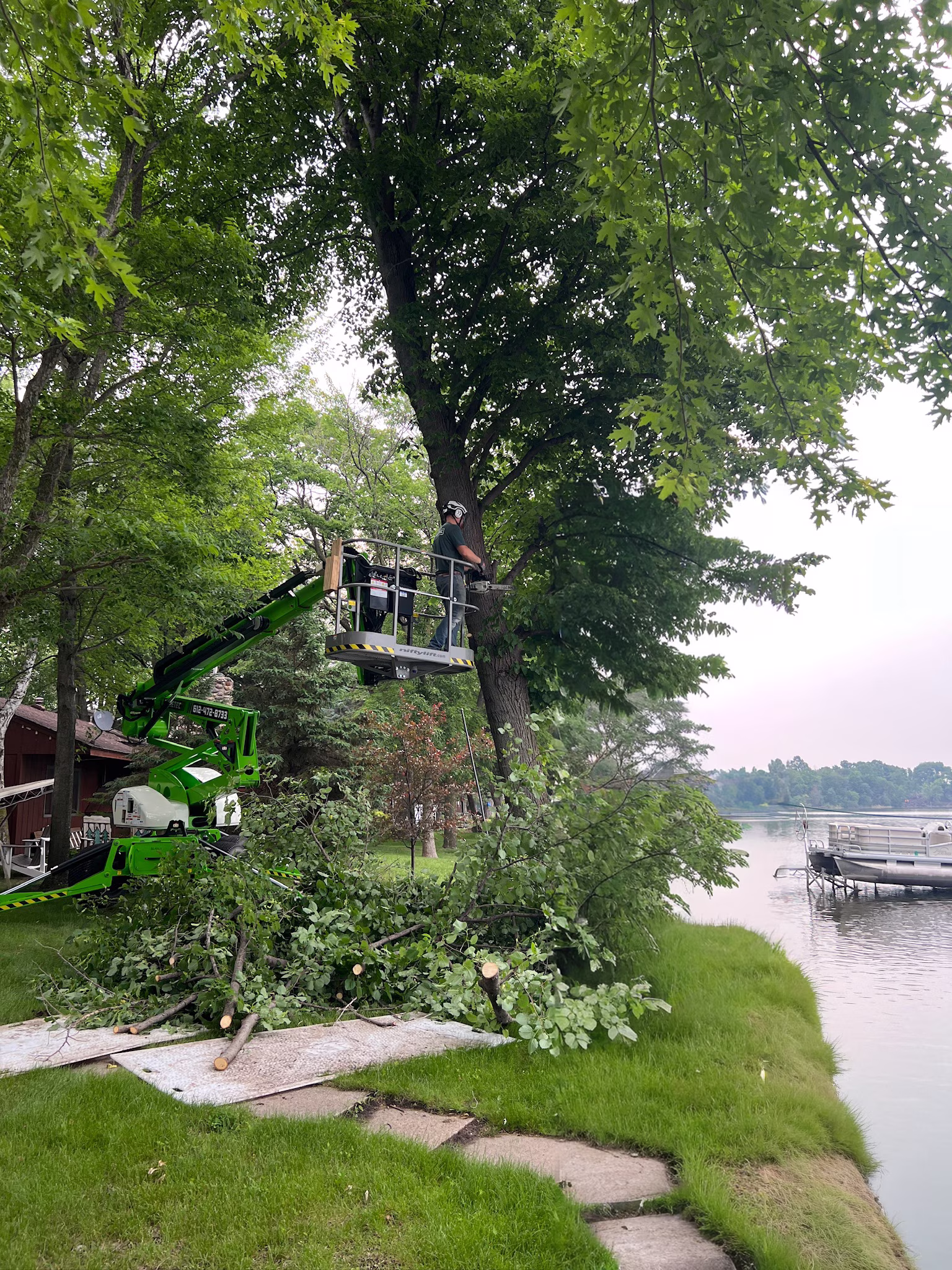 Worker in a lift cutting a large tree branch near a lakeside lawn with a docked boat visible in the background.