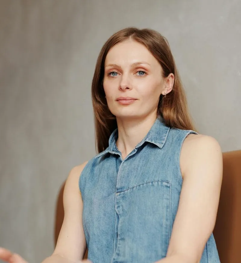 Serene woman with long brown hair wearing a sleeveless denim shirt sitting against a neutral background.