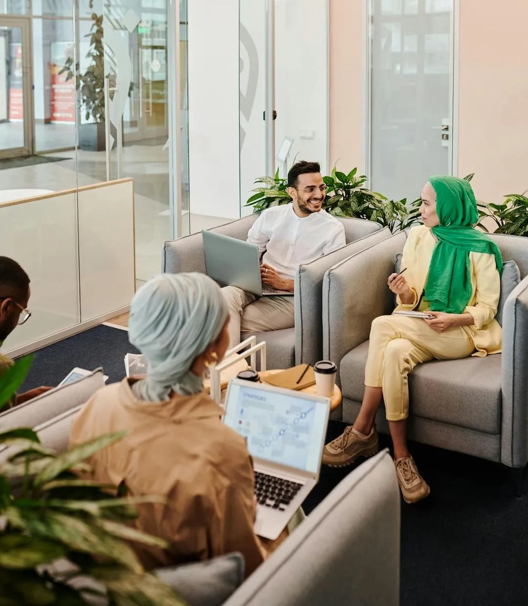 Four diverse professionals in a modern office lounge having a discussion with laptops and notebooks.