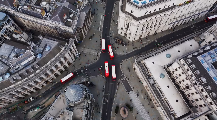 Aerial view of a city intersection with five red double-decker buses and surrounding historic buildings.