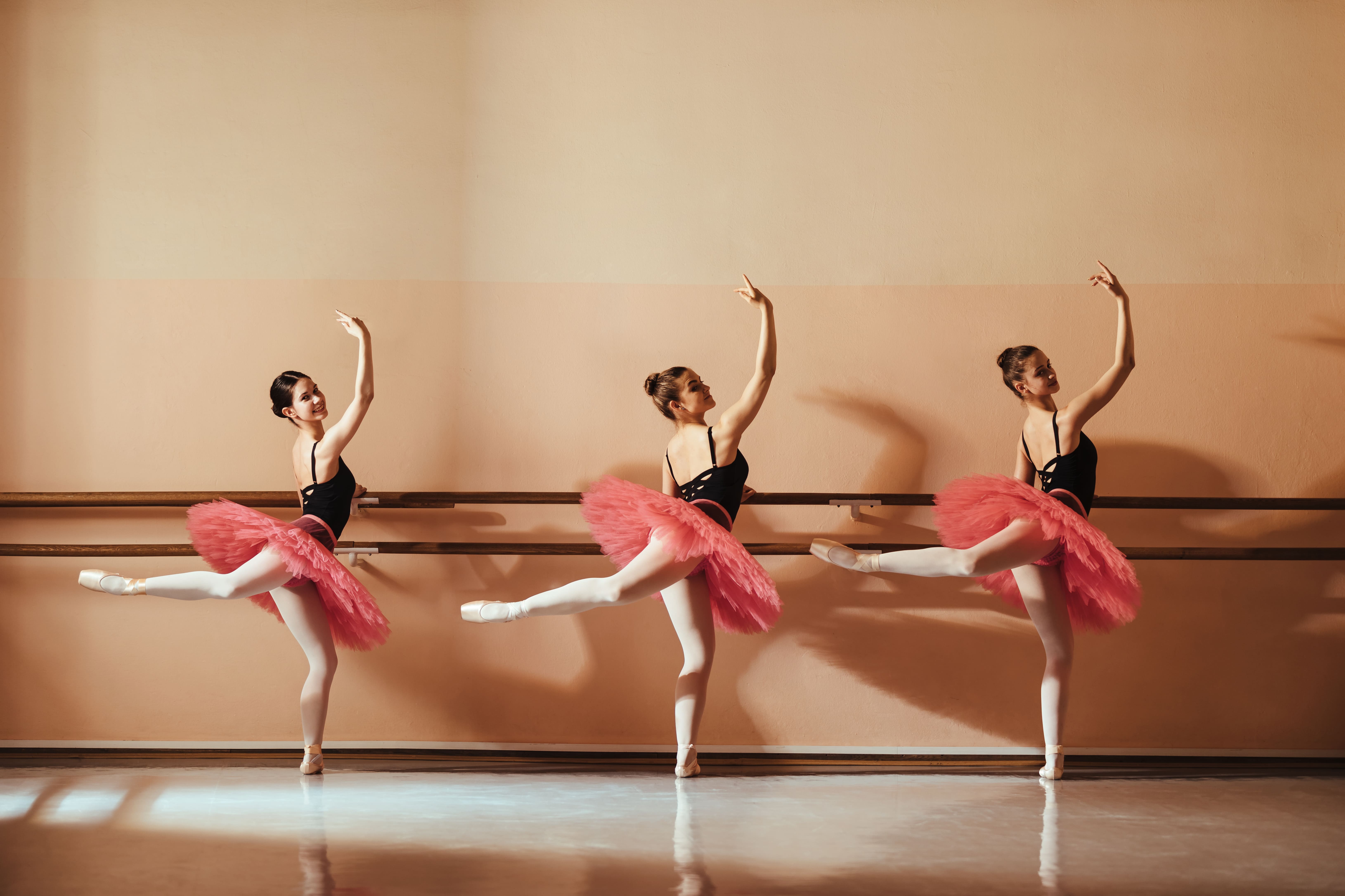 Three teenage ballerinas in black leotards and pink tutus practicing ballet at a barre, each with one leg raised behind.