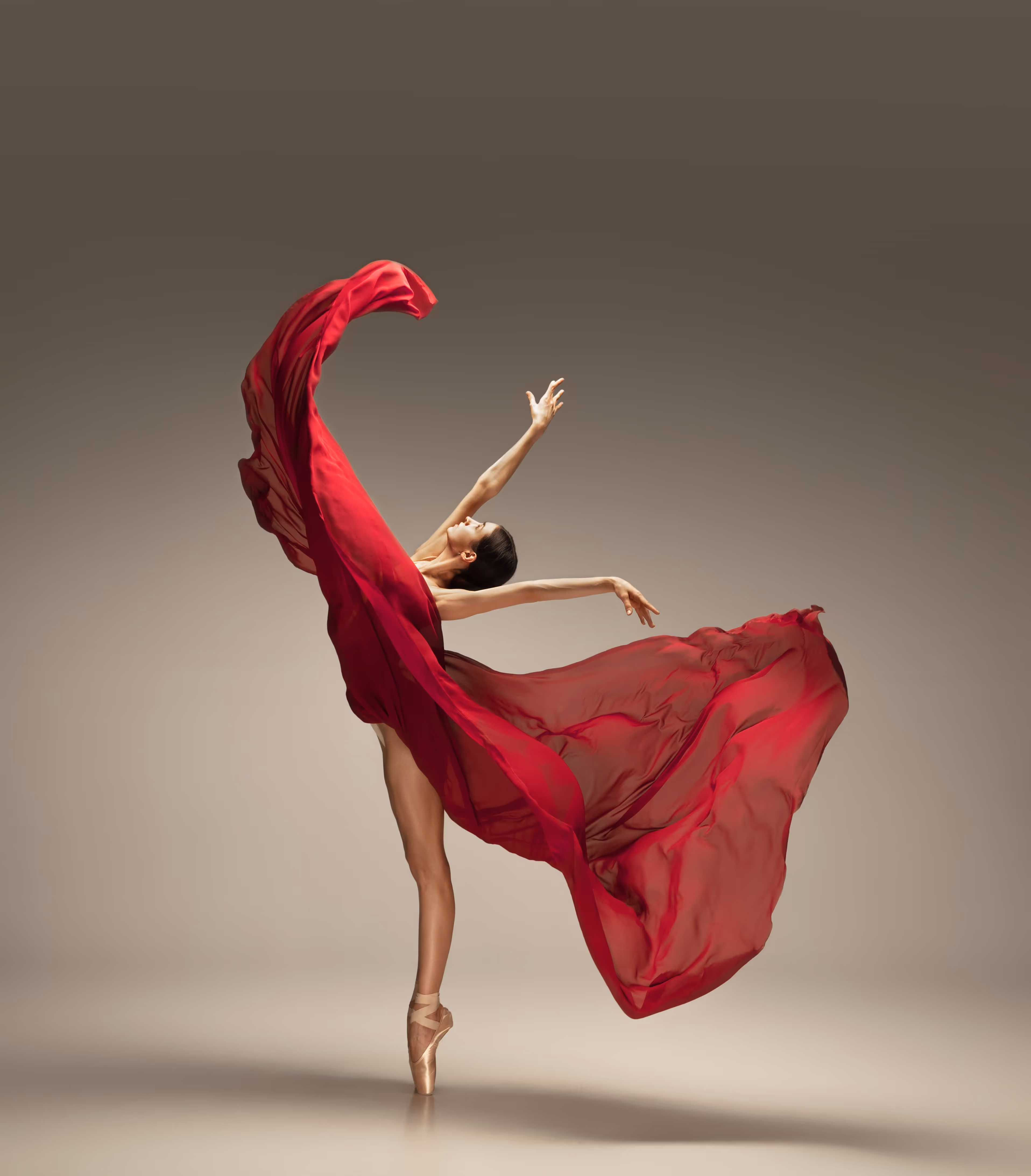 Ballerina on pointe with flowing red fabric arced gracefully in studio with neutral background.