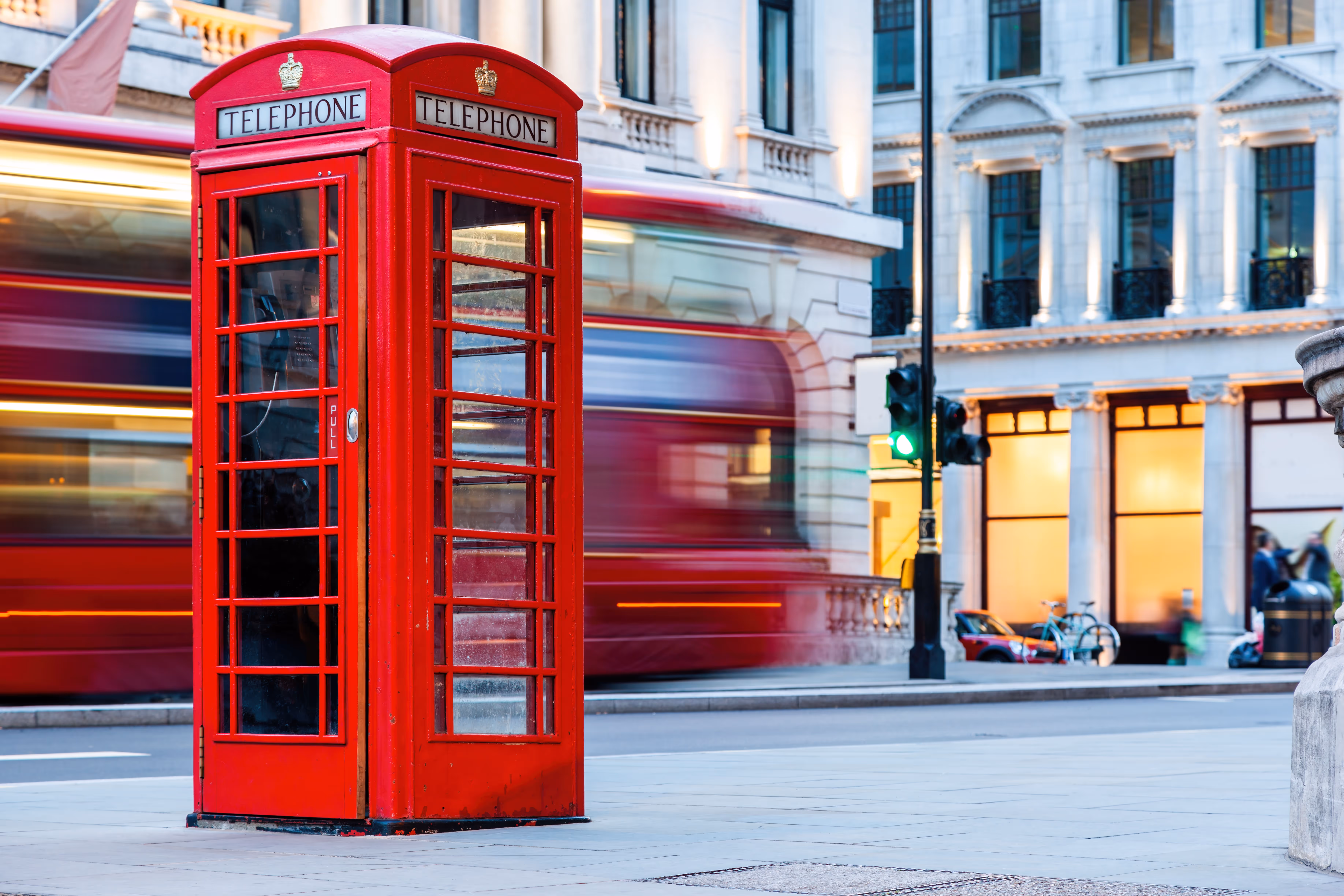 Classic red British telephone booth on a city street with a blurred red double-decker bus passing by at dusk.