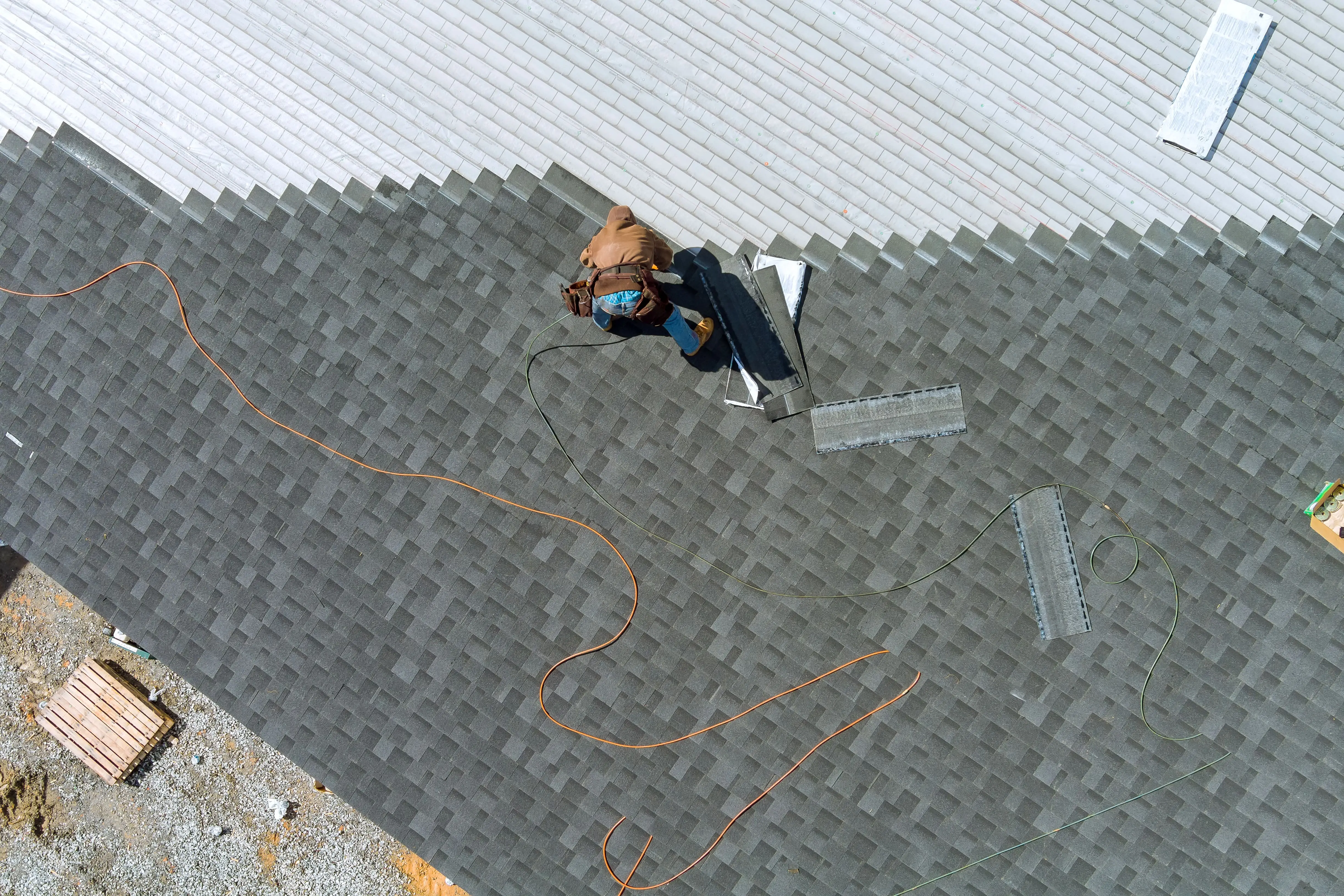 A roofer installs dark gray asphalt shingles on a roof, with tools and materials nearby and two long cords laid out.