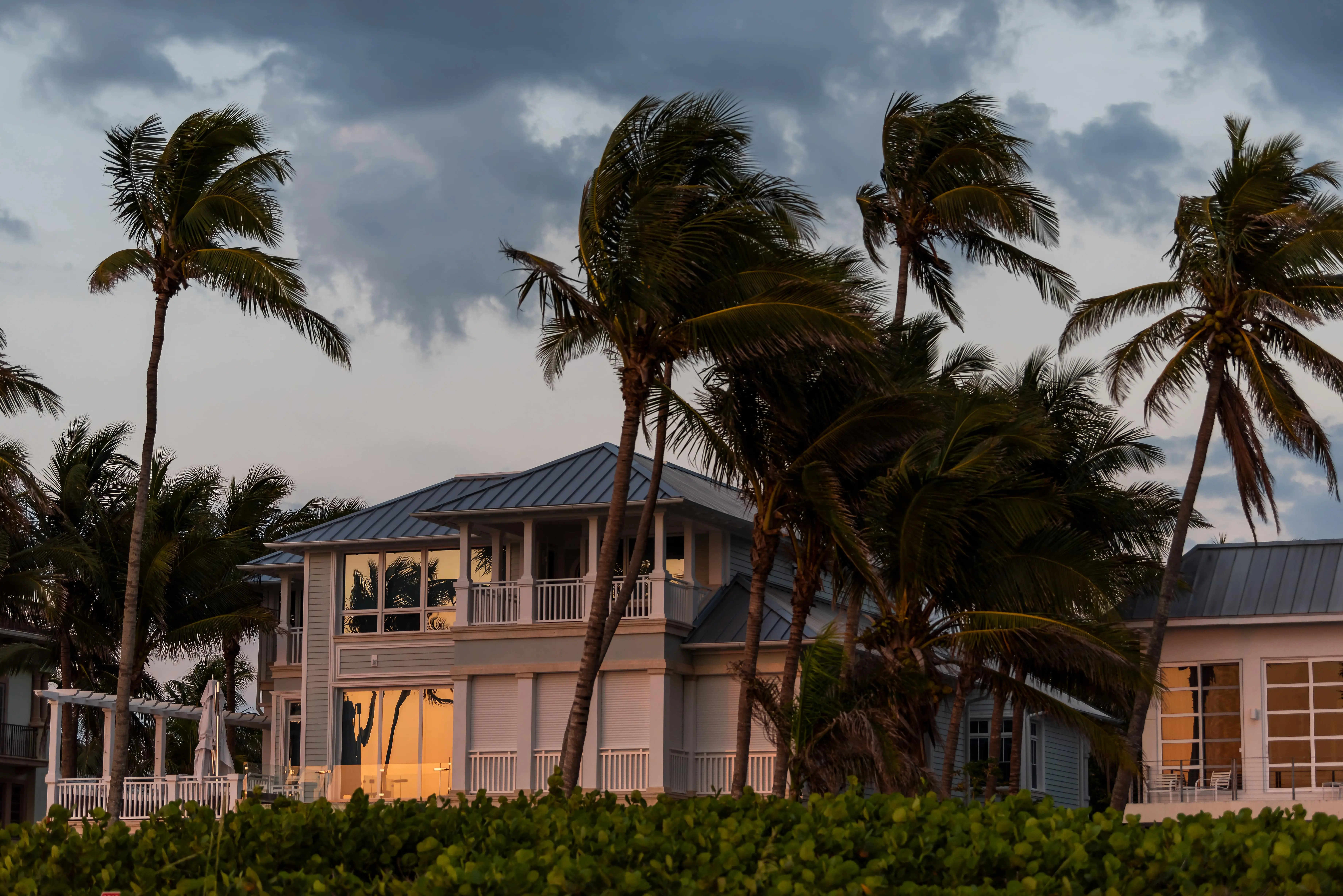 Modern two-story beach house with large windows reflecting a sunset, surrounded by palm trees and greenery.