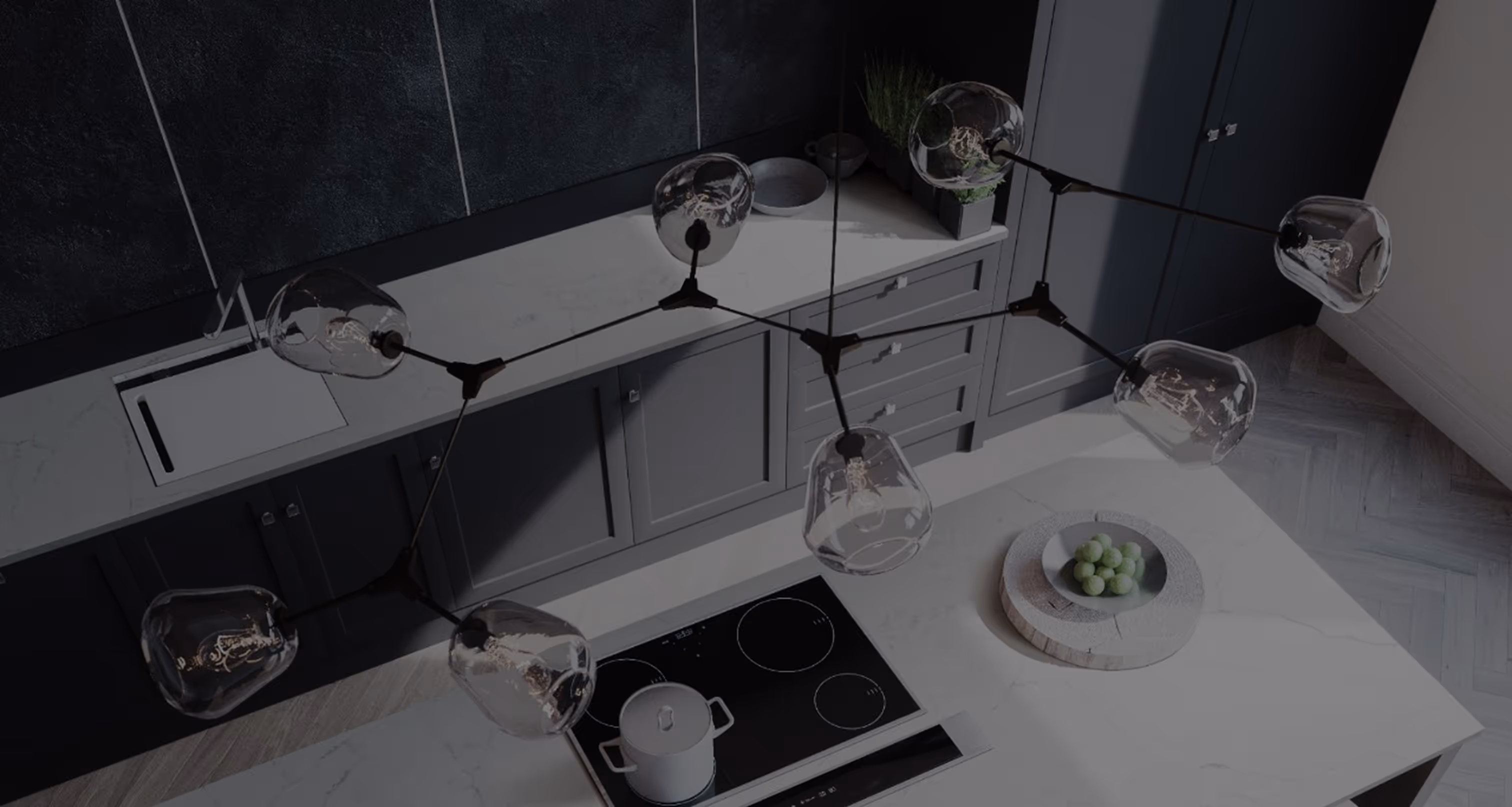 A modern kitchen featuring a unique overhead pendant light fixture with multiple bulb sockets. Below, a stovetop with a kettle and a white bowl holding green apples on a countertop. The cabinetry is dark gray.