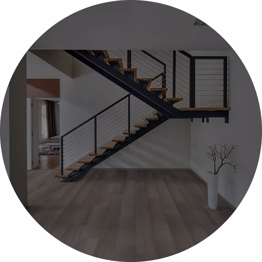 A modern interior featuring a staircase with wooden steps and black railing, leading to an upper level. The floor has light wooden planks, and a minimalist white vase with a branch is positioned to the right.