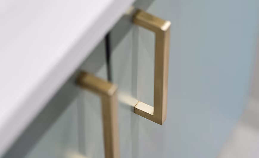 Close-up of modern cabinet handles with a minimalist design. The handles are gold in color and are attached to a light blue cabinet, reflecting a sleek and contemporary aesthetic.