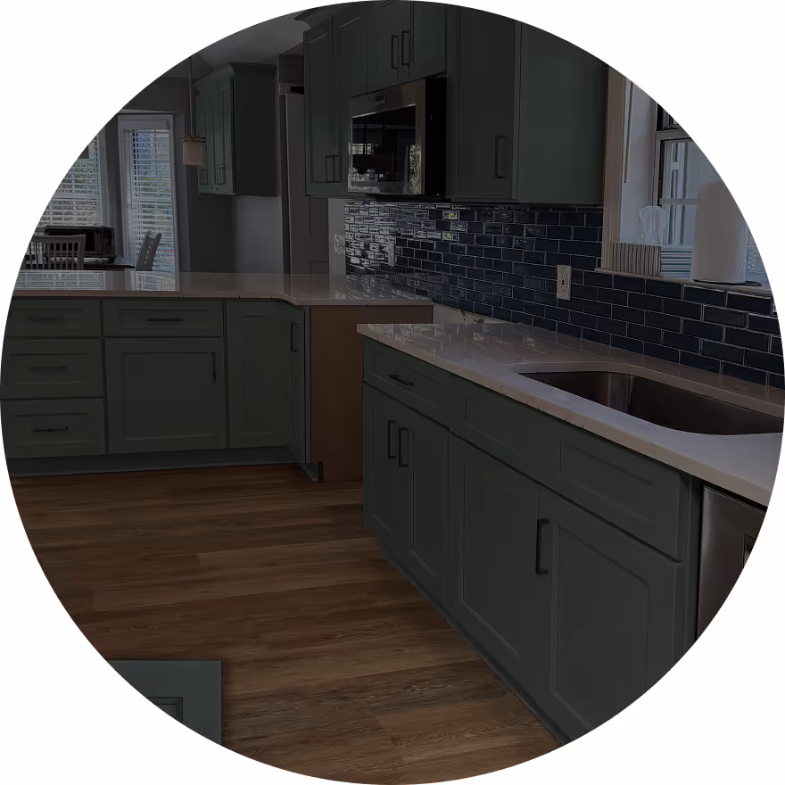 A modern kitchen featuring dark green cabinets, a light countertop, and a blue tile backsplash. The space has wooden flooring and natural light coming through a window.