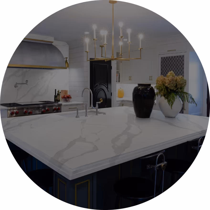 A modern kitchen featuring a large marble island with a sink, surrounded by high-backed stools. A black vase with flowers sits on one side, while a chandelier hangs overhead in a well-lit space.