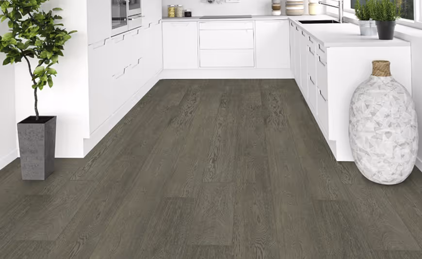 A modern kitchen featuring gray wooden flooring, white cabinets, and a large window with potted plants. A decorative vase sits on the counter, contributing to the contemporary design.