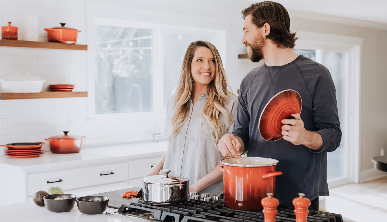 A couple smiling at each other while cooking in a modern kitchen. The man is stirring a pot on the stove and holding a lid, while the woman stands beside him. Cookware is arranged on the counter.